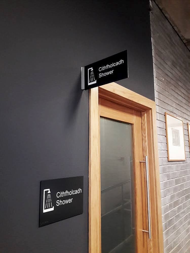 Bilingual shower sign in the UCD Tierney Building: white shower pictogram on a black panel with Cithfholcadh and Shower in Irish and English, flag-mounted above the door and repeated at hand height