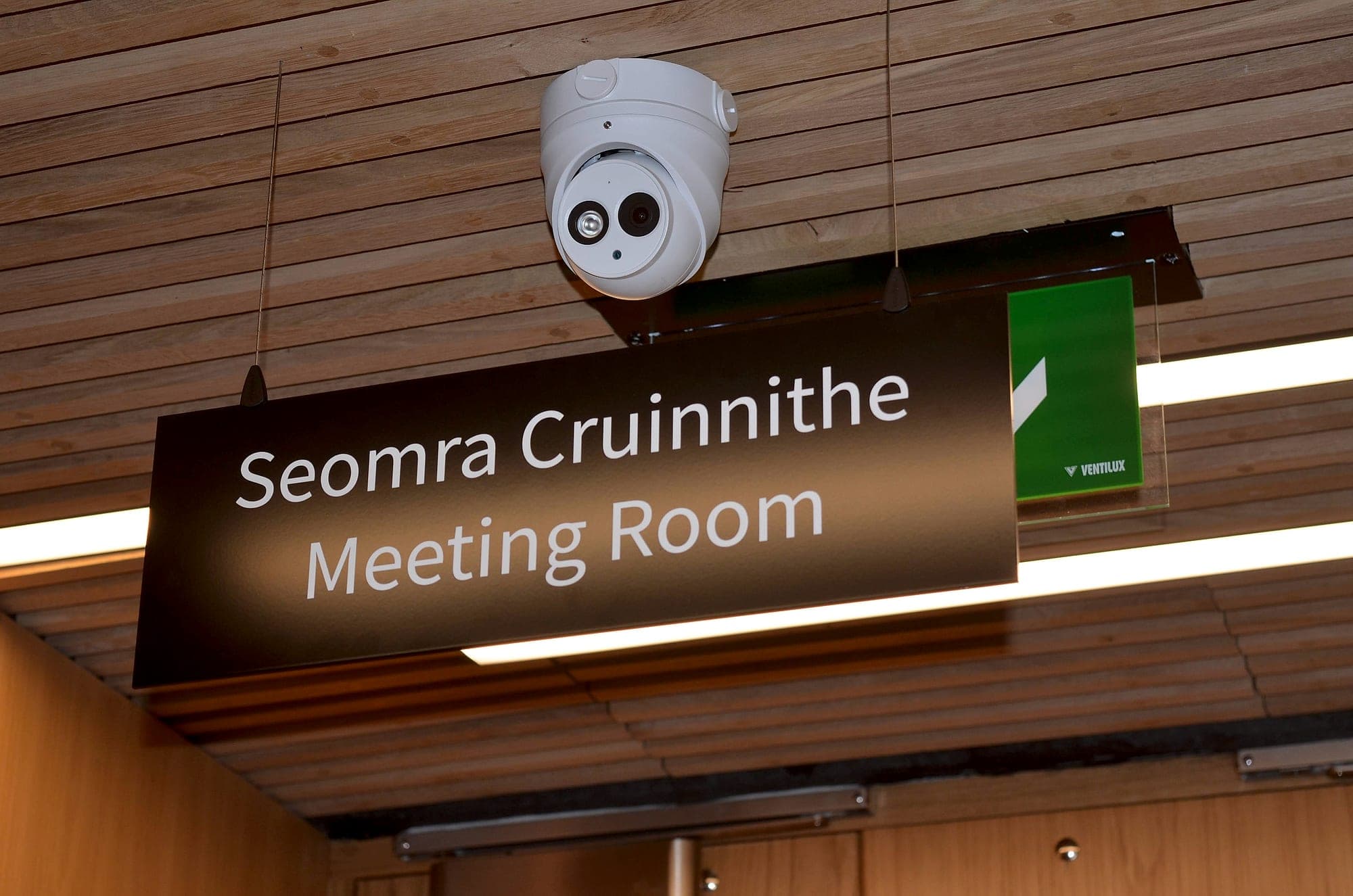 The bottom of the pyramid: bilingual Irish and English suspended sign reading Seomra Cruinnithe / Meeting Room, hung from a timber-slatted ceiling above a doorway