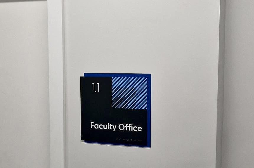 Braille signage beside a room door at CCT College Dublin: a tactile identification sign with raised room number, raised mixed-case name and a braille line below, mounted on the wall on the latch side