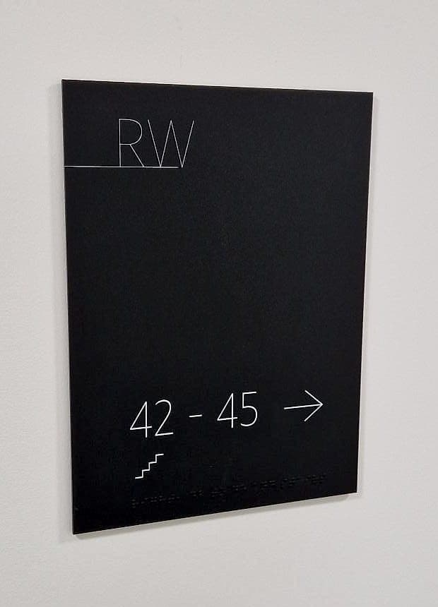 Braille signage on an apartment corridor wall at Rathborne Wharf, Dublin: matt black panel with white flat numbers, a stair pictogram and a raised braille line beneath