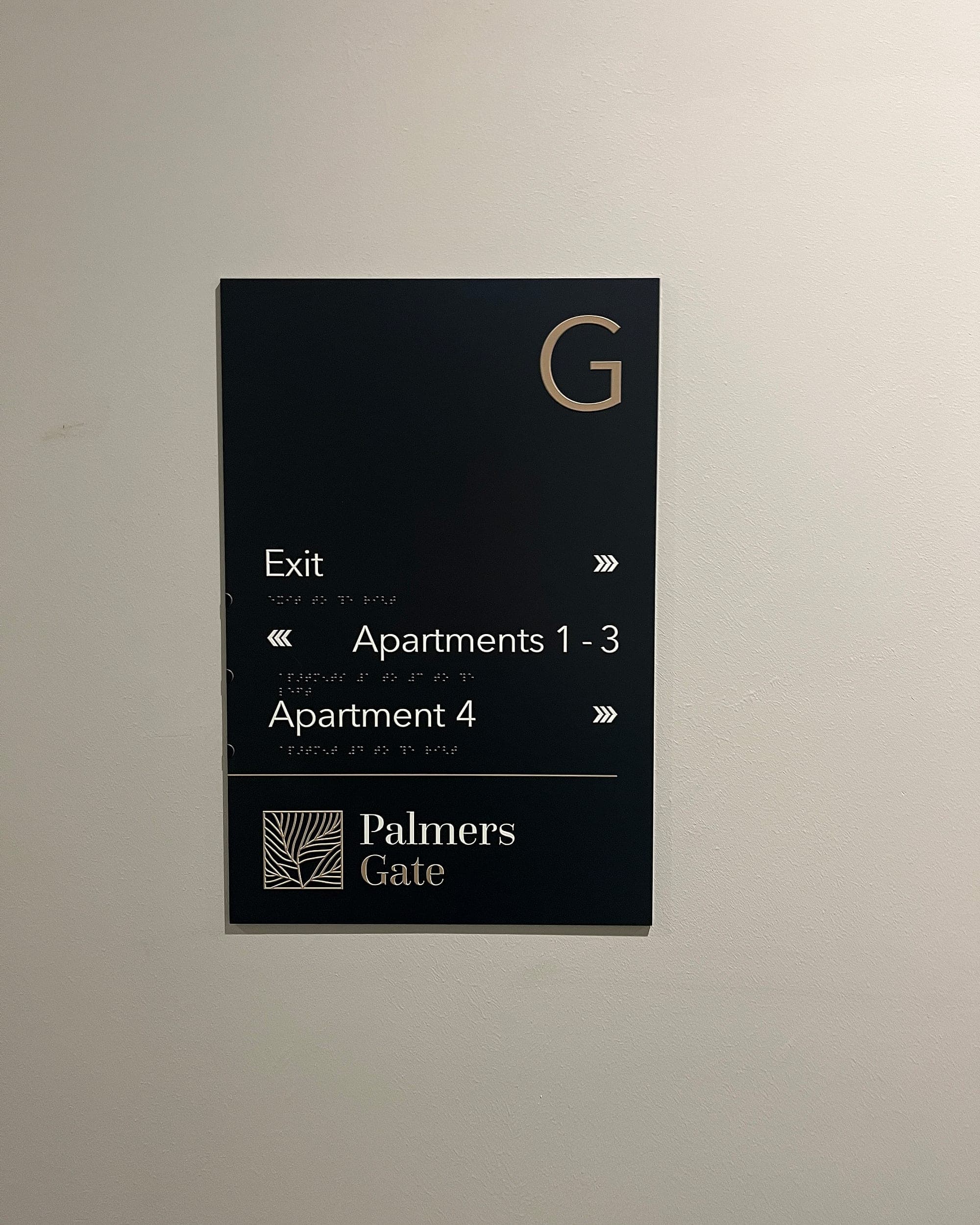 black ground-floor directional panel at Palmers Gate with white lettering for Exit and apartment ranges, gold chevron arrows and a raised braille line set beneath every destination
