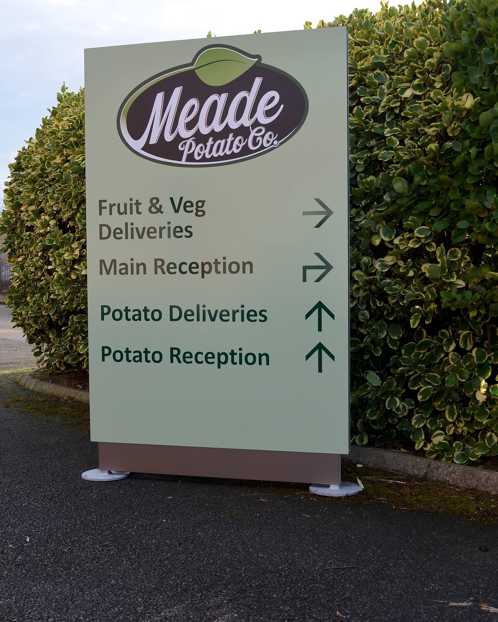Meade Potato Company, site entrance totem for a food producer: brand logo on a pale sage brand-tint field above directional lines set in mid-brown and deep green with arrows