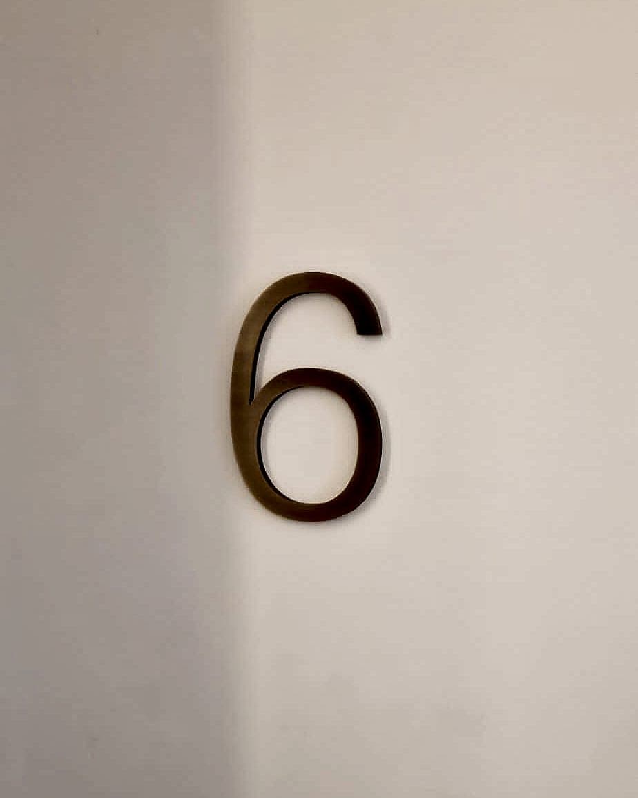 Solid antique-brass numeral six at Leeson Place, face-fixed directly to a plain wall in a residential circulation space, dimensional lettering used instead of a printed panel