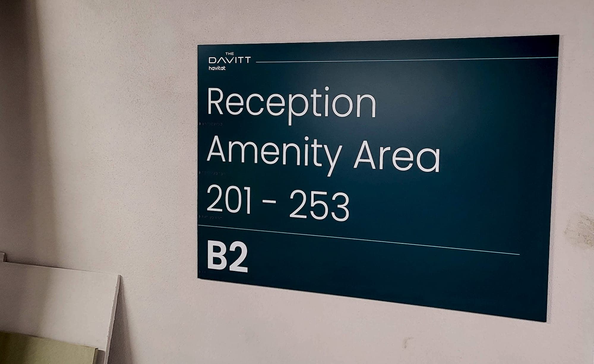 Build-to-rent amenity signage: a teal directional panel in the operator's own brand naming Reception and Amenity Area with apartment numbers 201–253 and level B2, with braille beneath each line