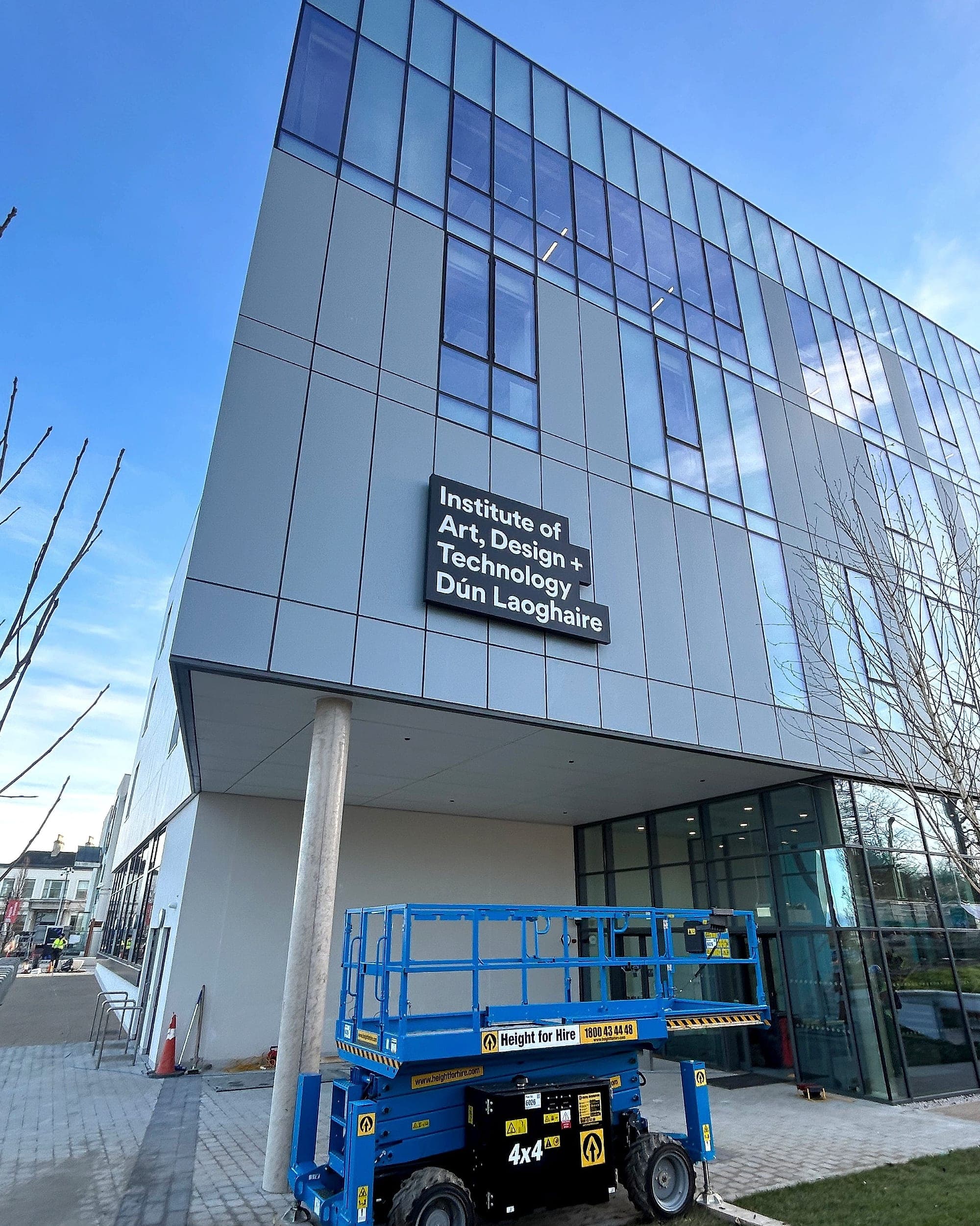 Building identification signage installed high on the façade of a new academic building at IADT Dún Laoghaire, with a scissor lift parked at the entrance below
