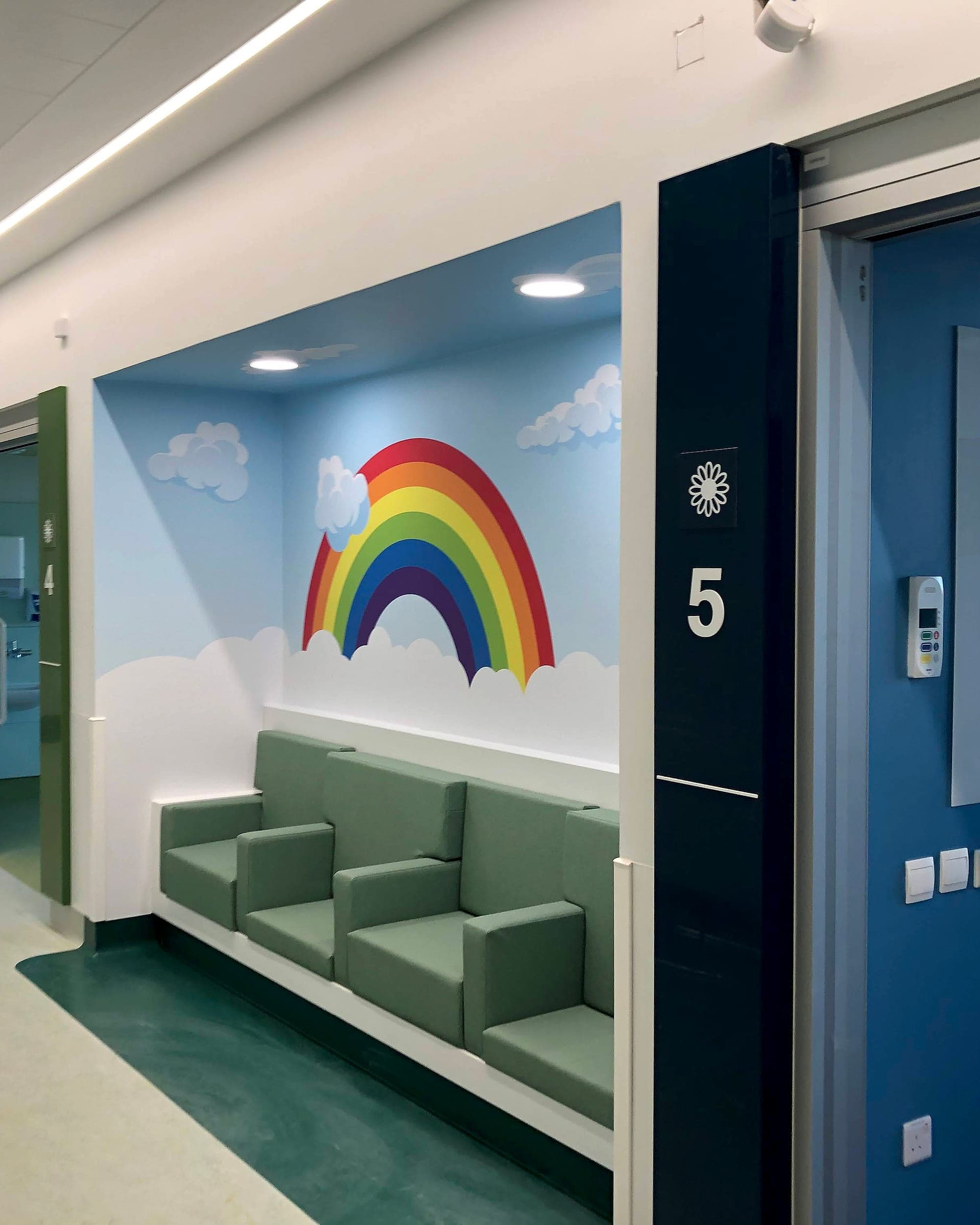 The National Rehabilitation Hospital: calm rainbow mural above a seating bay in a ward corridor, beside a room number sign carrying its own symbol. Autism friendly signage and landmark together