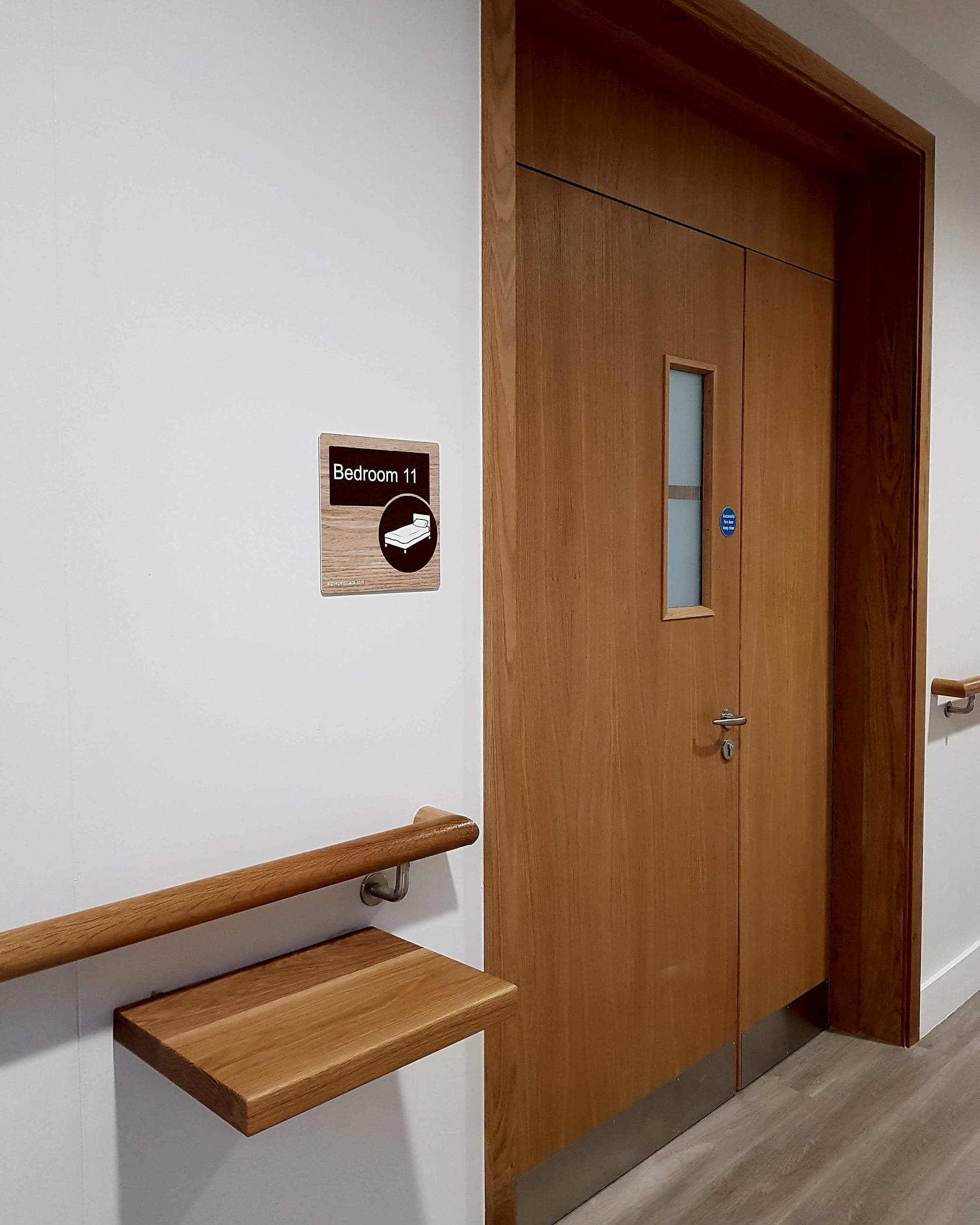 Care home bedroom door sign mounted at hand height on the latch side, with a bed pictogram, beside an oak handrail and a rest shelf