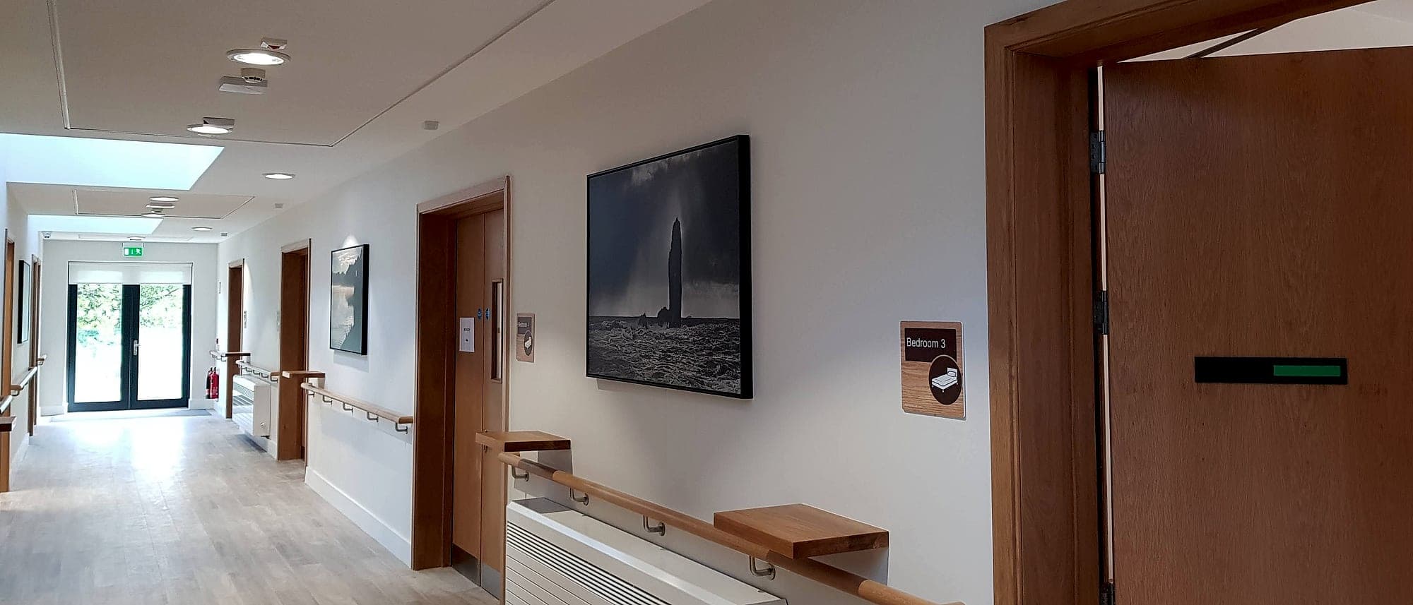 Care home corridor using framed photographs as landmarks, with an oak handrail and rest shelf and a bedroom door sign carrying a bed pictogram
