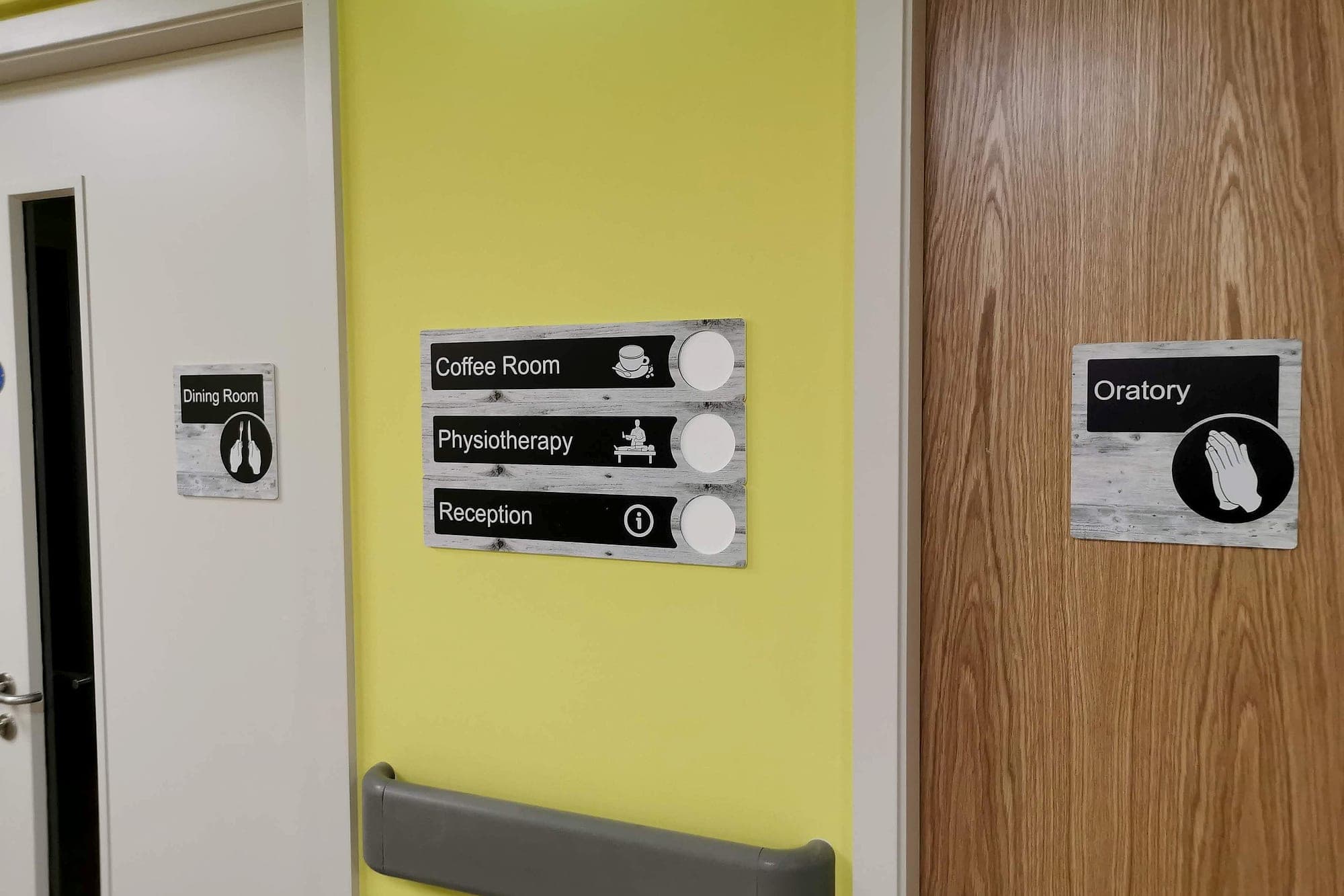 Dementia-friendly pictogram signs for coffee room, physiotherapy and reception mounted on a yellow accent wall, with an oratory door sign and a dining room identification sign alongside