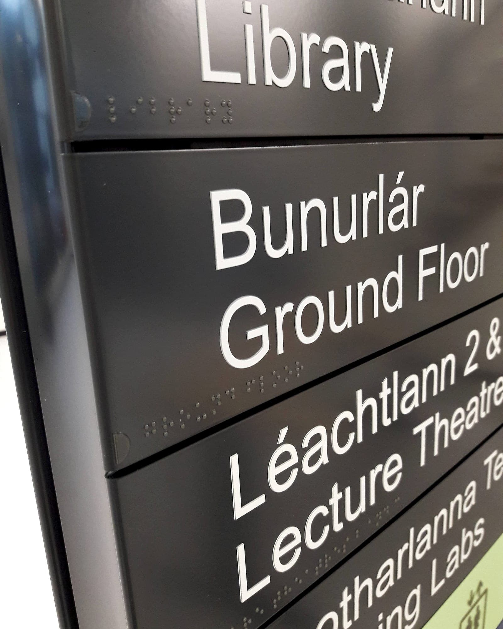 Close-up of a modular slat directory: separate removable aluminium slats carrying bilingual Irish and English text with braille beneath each line