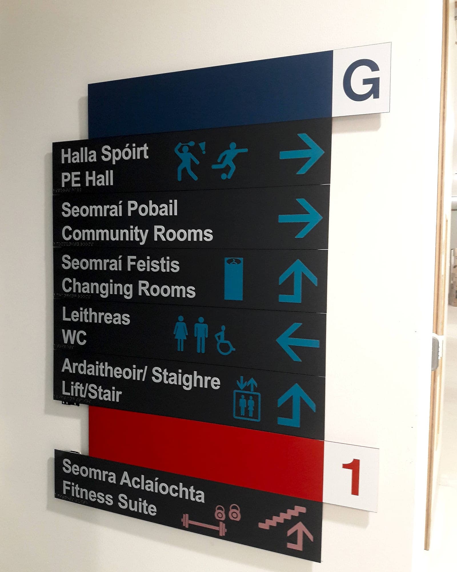 Bilingual Irish and English directional signage at Adamstown Community Centre with colour-coded floor bands, blue for ground level and red for level one, with high-contrast pictograms and arrows
