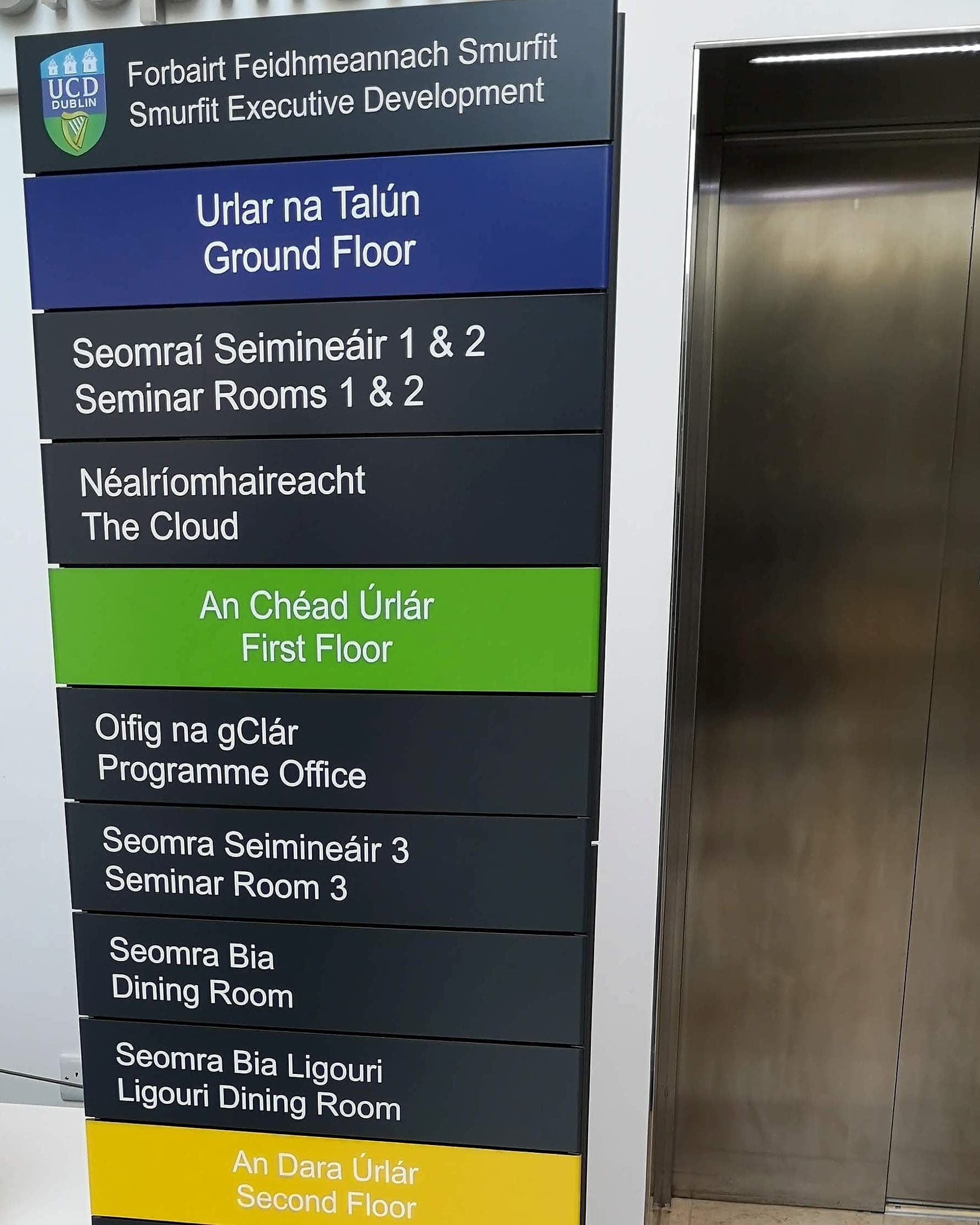 Colour-coded floor bands on a bilingual internal directory at UCD Smurfit Executive Development: blue for the ground floor, green for the first floor, yellow for the second, each colour also named in Irish and English