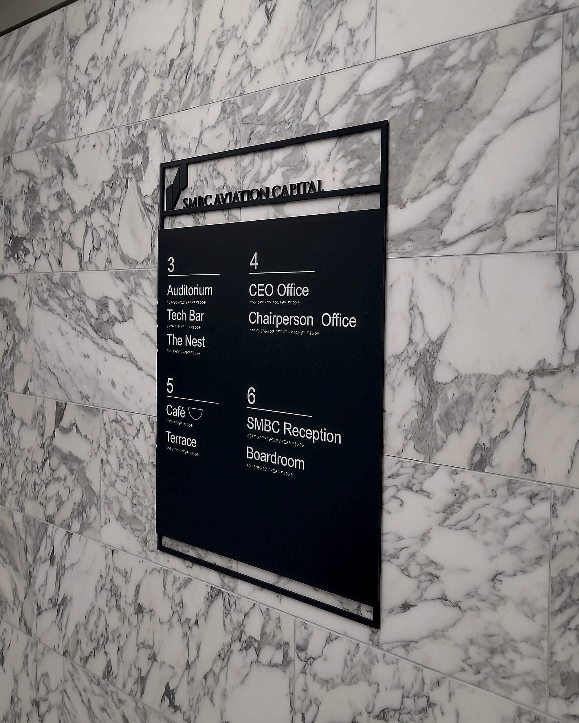 Corporate internal directory sign at SMBC Aviation Capital on a marble wall, listing floors 3 to 6 with destinations including Auditorium, CEO Office, Café, Reception and Boardroom in white lettering with braille beneath each line