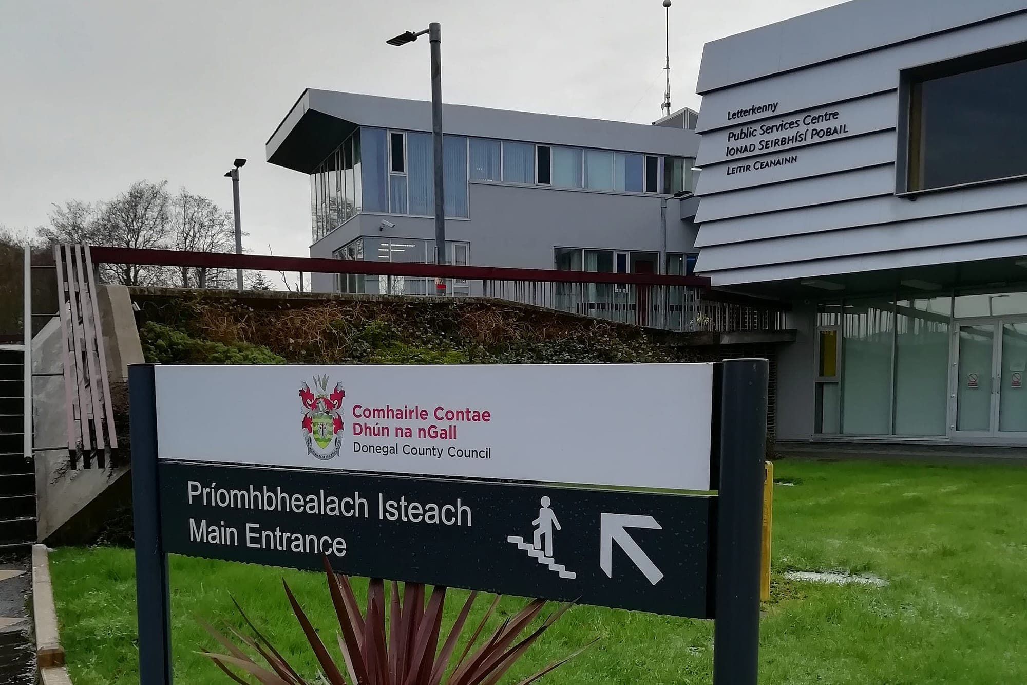 Bilingual Irish and English post-and-panel main entrance sign with the county crest at Donegal County Council's Letterkenny Public Services Centre, with the building's own bilingual lettering on the facade behind