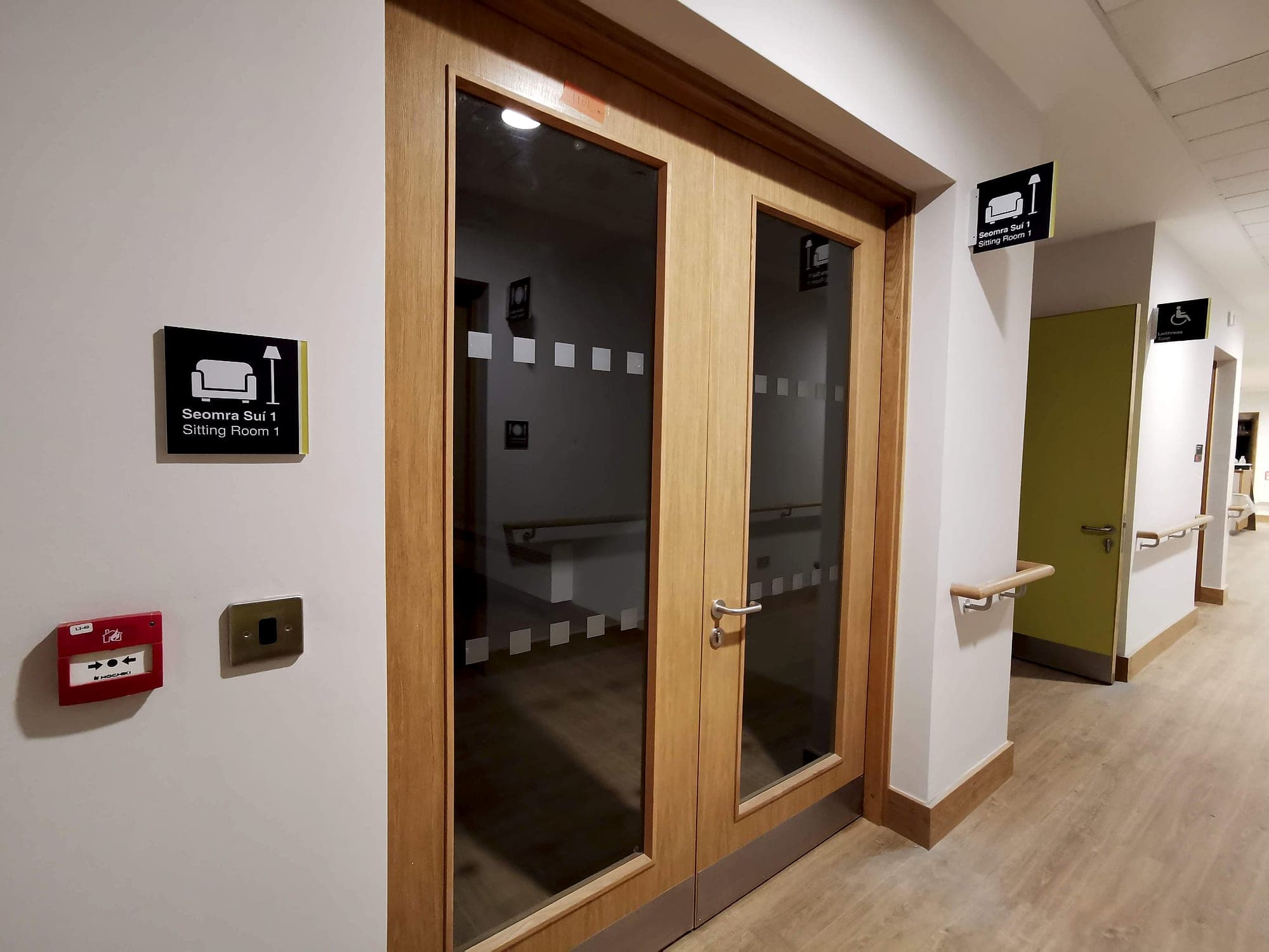 Custom wayfinding pictogram of an armchair and standard lamp on a bilingual door sign reading Seomra Suí 1 and Sitting Room 1, repeated on a flag-mounted projecting sign along the corridor with an accessible toilet symbol beyond
