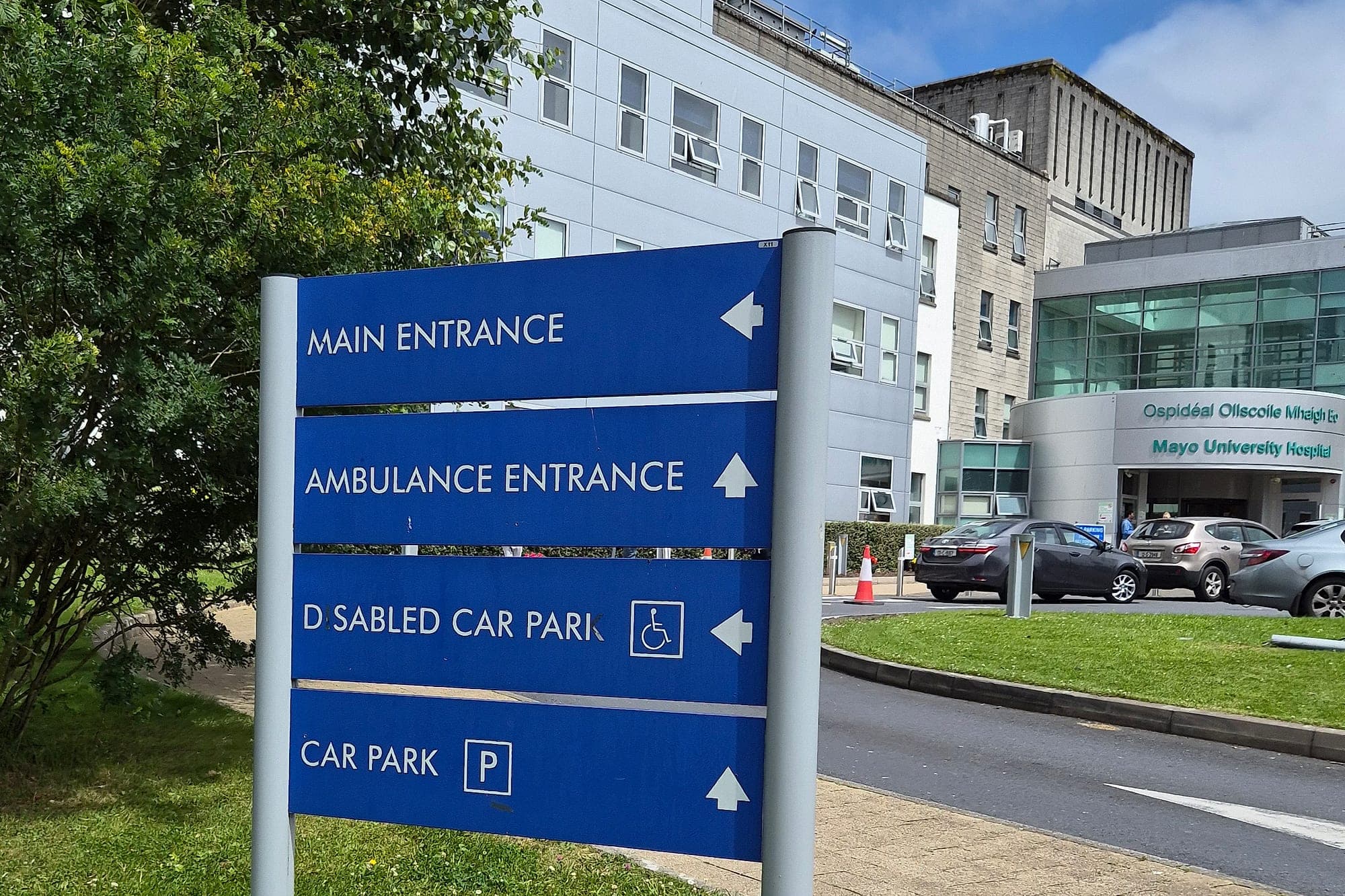 The finding that opened the survey (accessibility audit finding): weathered blue hospital directional sign set entirely in capitals, with a damaged legend where the accessible car park is signed alongside the International Symbol of Access