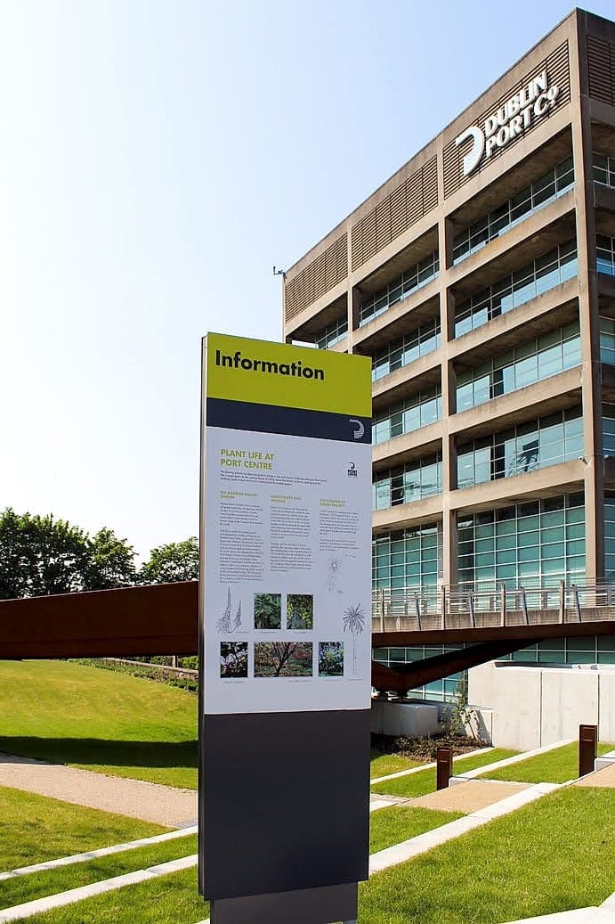 Information monolith at Dublin Port with black text on a lime-green header panel and a white information panel on a dark grey totem, the Port Centre building behind