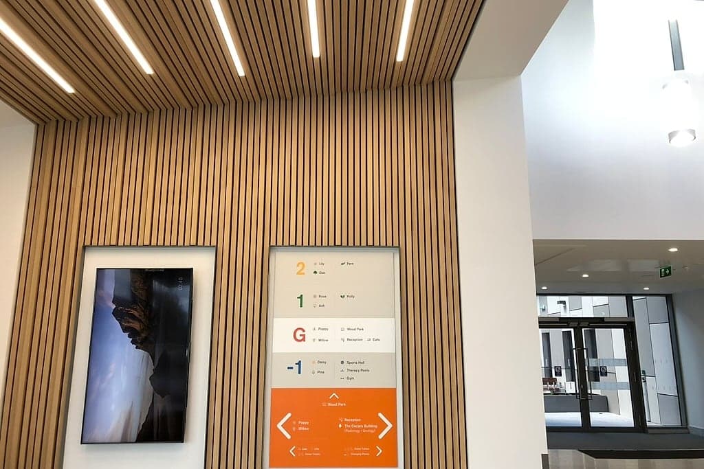 Smart building signage in the lobby at the National Rehabilitation Hospital: a wall-mounted digital display beside a fabricated floor directory listing wards and departments by level, both recessed into a slatted timber wall