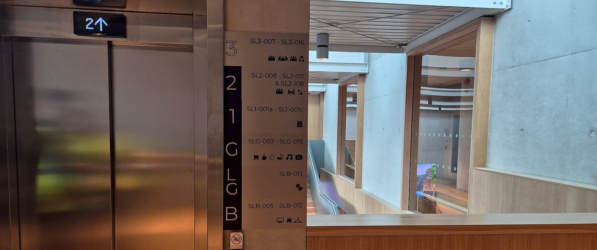 Digital wayfinding and static signage in one lift lobby: a fabricated floor directory strip beside the lift doors, with a digital display visible across the atrium beyond