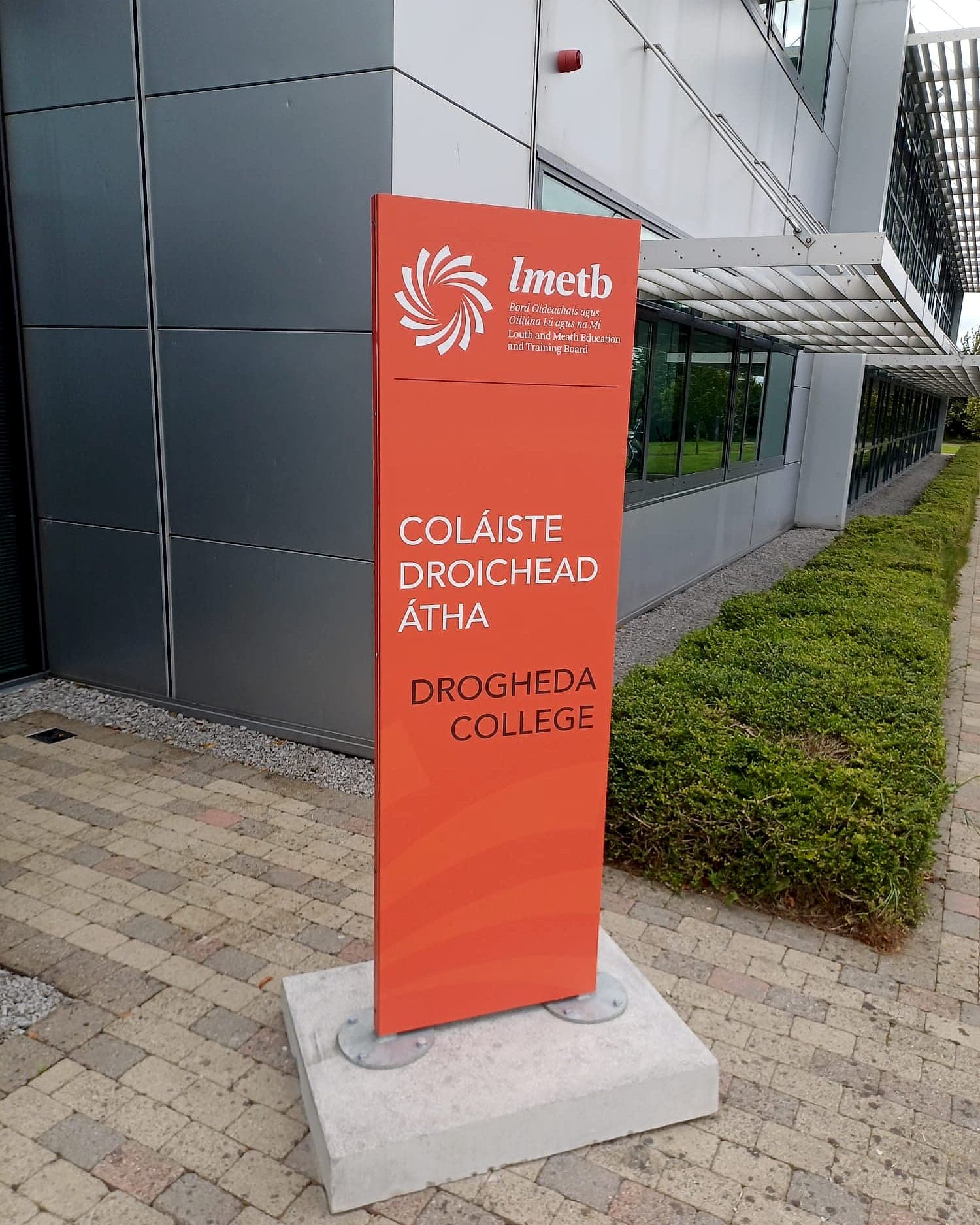 Bilingual orange entrance identity totem reading Coláiste Droichead Átha / Drogheda College with the LMETB mark, bolted through flanged feet to a concrete pad set into block paving beside a clad building entrance