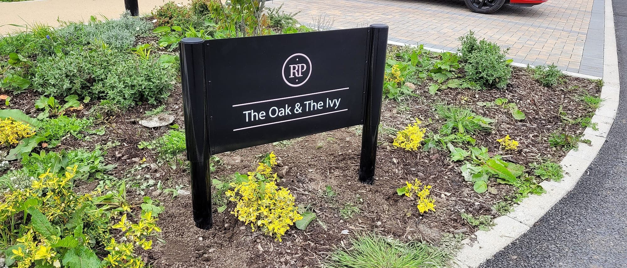 Black post-and-panel building identification sign at Rathyvoy, Naas, set in a planting bed at the edge of a block-paved drive