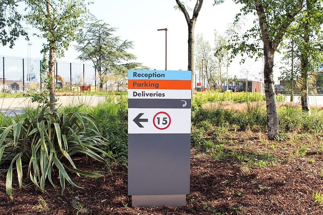 External directional monolith totem at Dublin Port with colour-banded panels for reception, parking and deliveries, an arrow and a 15 km/h speed roundel