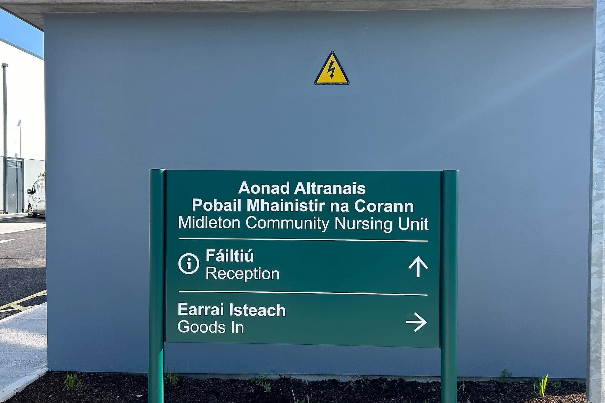 External bilingual Irish and English directional sign for Midleton Community Nursing Unit in front of a plant enclosure carrying a yellow electrical hazard warning triangle