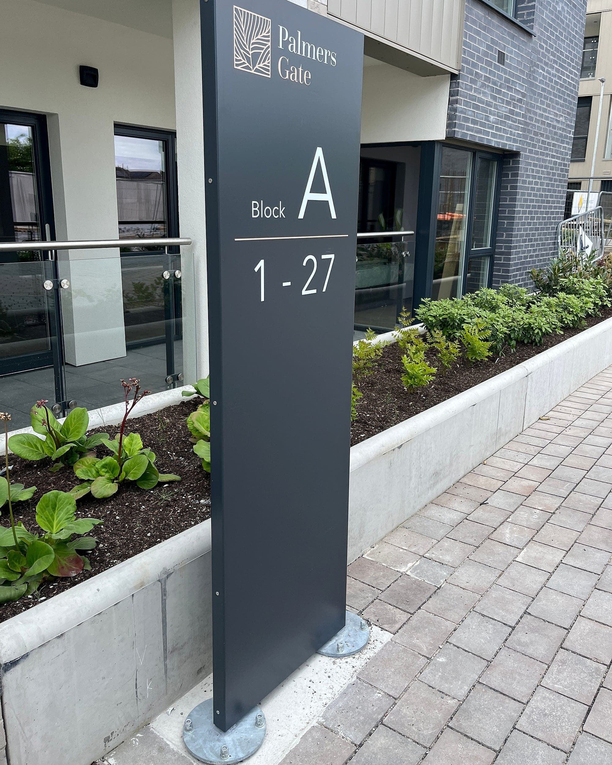 External signage foundations and fixings: dark grey aluminium block identification totem at Palmers Gate residential scheme, bolted down through paving on two galvanised baseplates