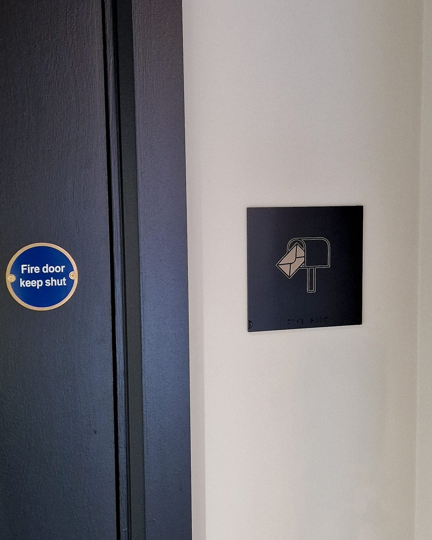 Blue circular Fire door keep shut safety sign in a brass ring on a navy door, beside a navy tactile and braille post-room pictogram sign: a statutory safety sign specified to the same quality as the building's wayfinding