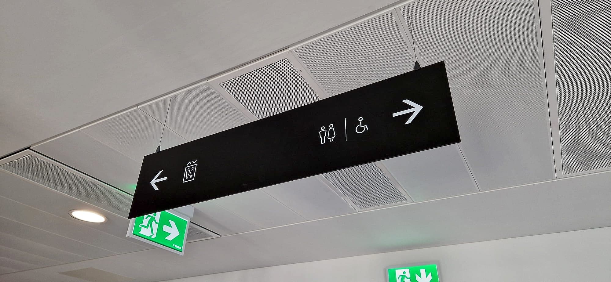 Fire safety signage in Ireland: a green illuminated running-man emergency exit sign mounted directly beneath a black suspended wayfinding blade in a corridor