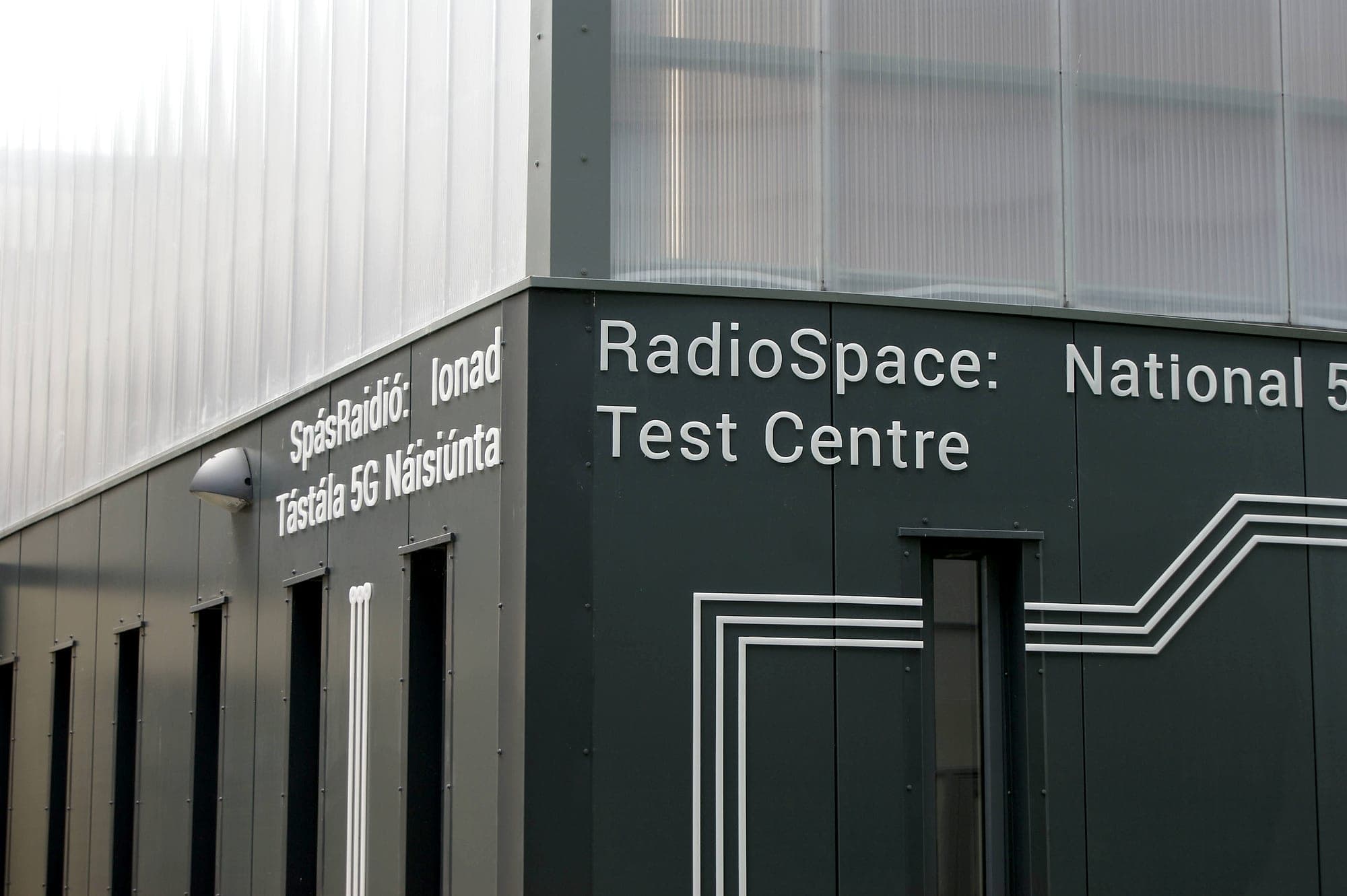 Bilingual Irish and English building identification signage at Maynooth University: flat-cut lettering face-fixed to dark grey composite cladding panels on an external wall