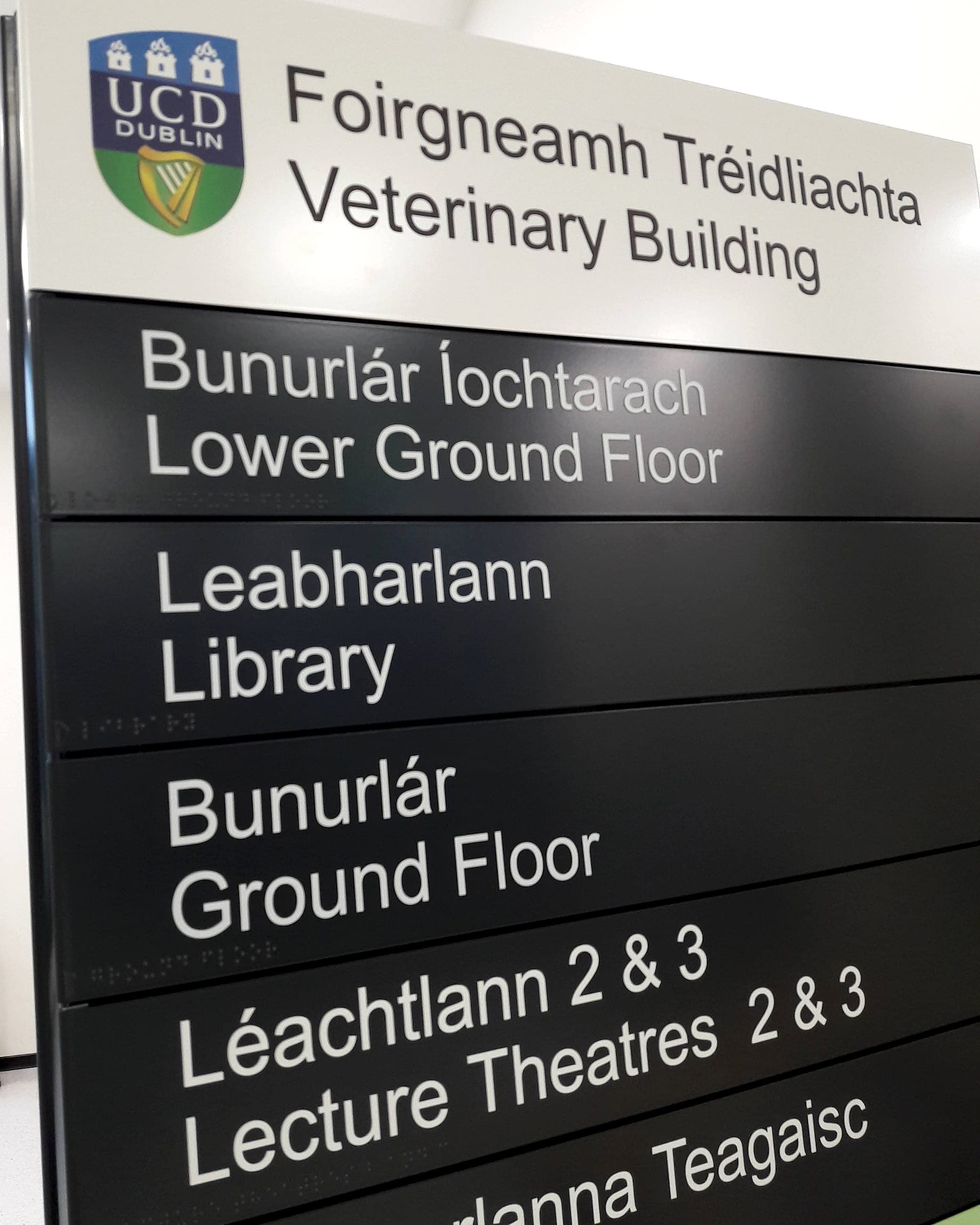 Bilingual Irish and English floor-by-floor directory sign at the UCD Veterinary Building, listing Lower Ground Floor, Library, Ground Floor and Lecture Theatres with braille beneath each line