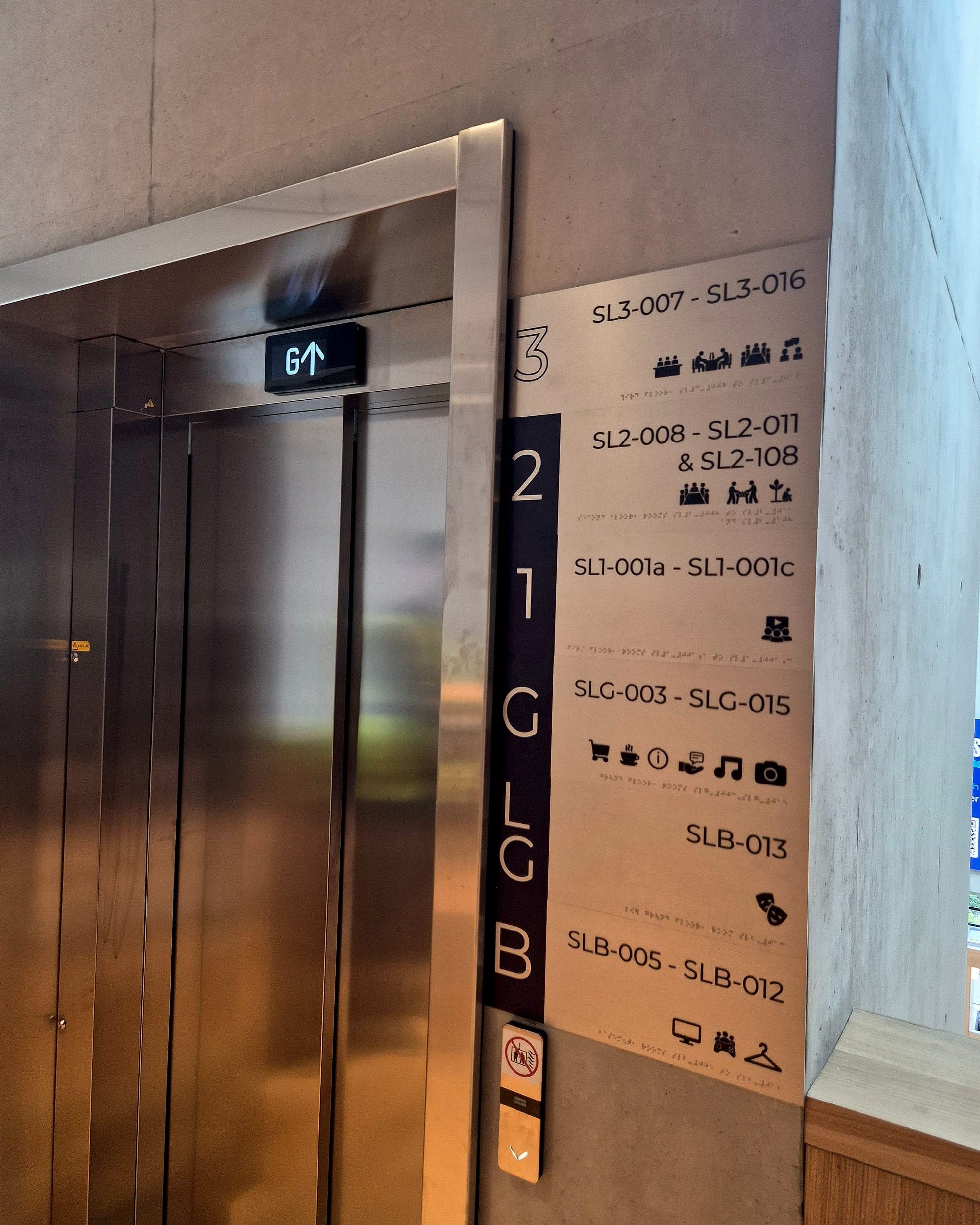 Floor directory sign beside a lift at the Glucksman Library, listing room-code ranges by level (SL3-007 to SL3-016, SLG-003 to SLG-015), with pictograms and braille beneath each line