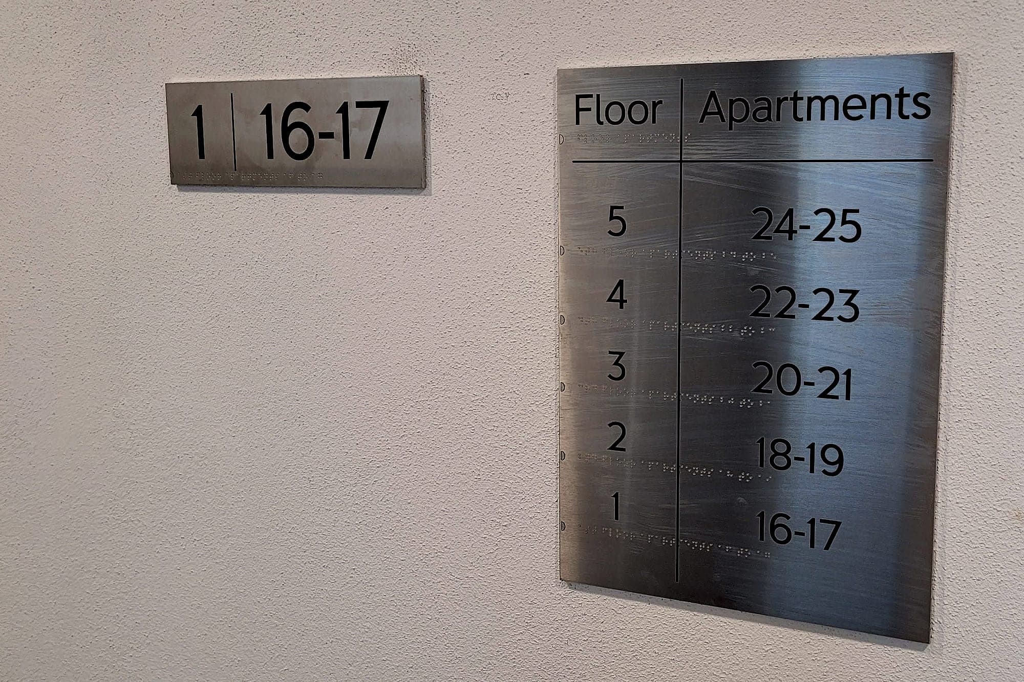 Floor identification and flat indicator signs in an apartment block lift lobby: a brushed stainless plate reading 1 | 16-17 beside a Floor / Apartments board listing floors one to five against apartment ranges
