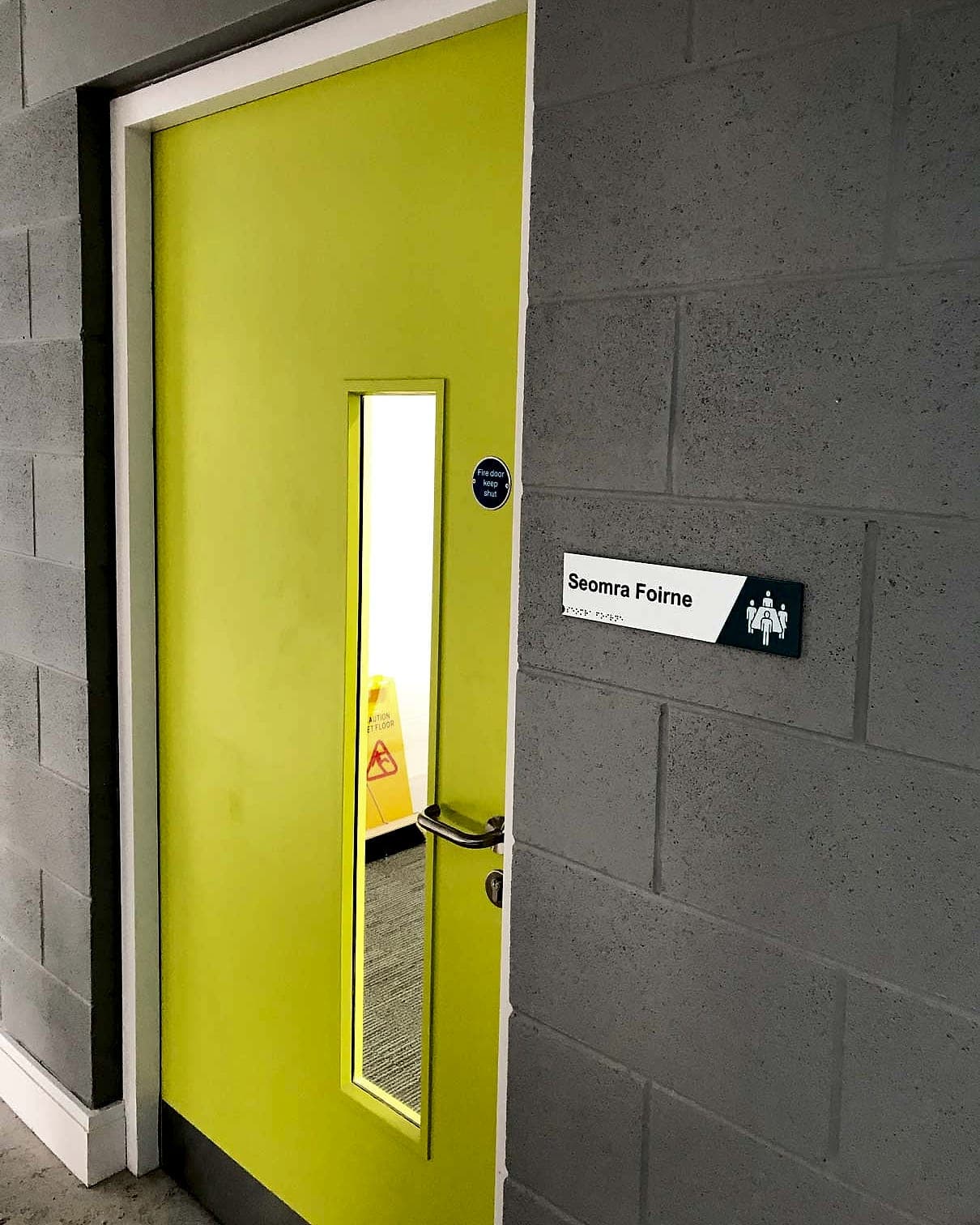 Irish-language door sign at Gaelscoil Bhaile Munna reading Seomra Foirne with raised tactile lettering, braille and a staff pictogram, mounted on the latch side of a fire door