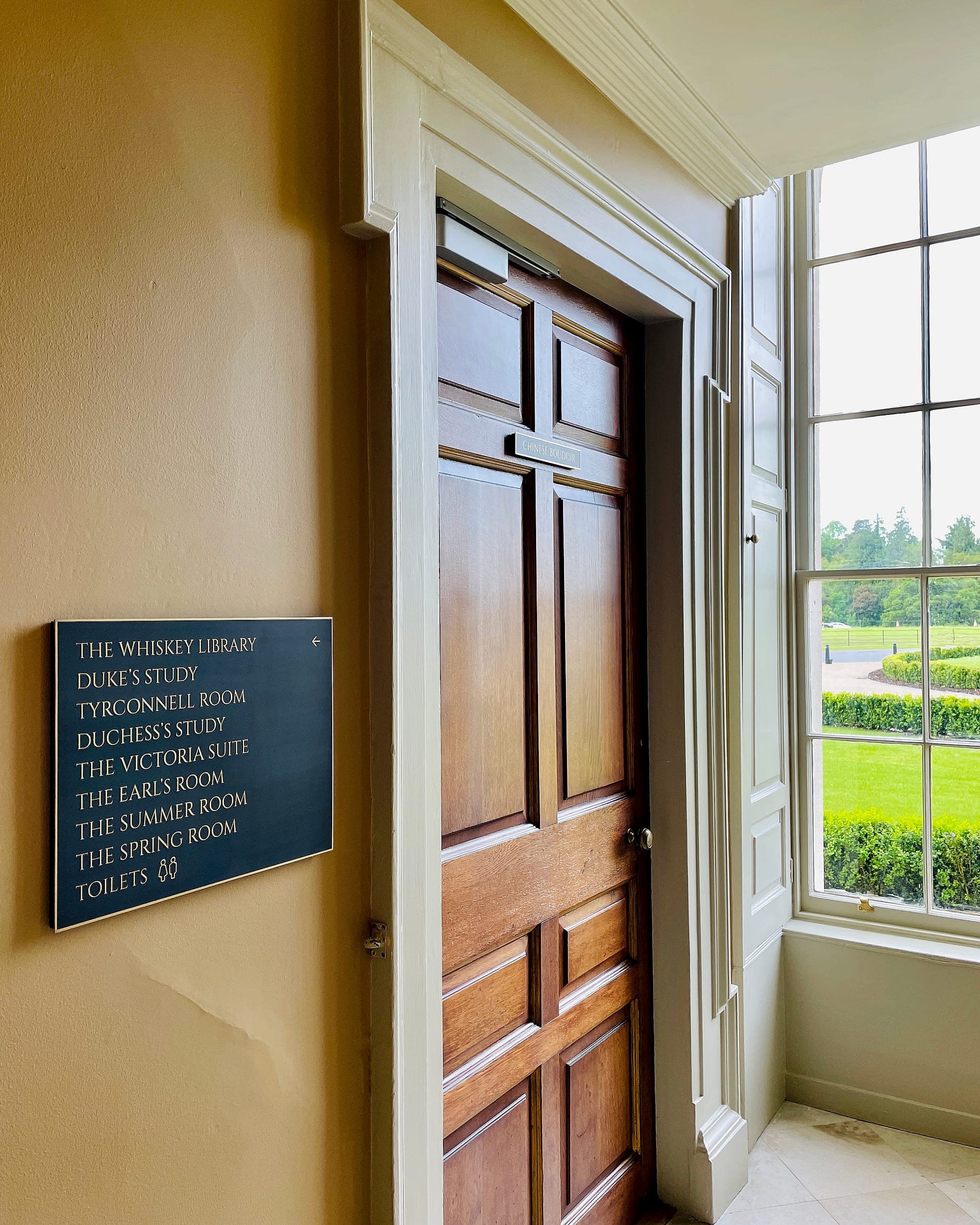 Heritage interior signage at Carton House: a dark blue directory panel with gilded lettering and a brass border, mounted beside a panelled oak door in a Georgian hall