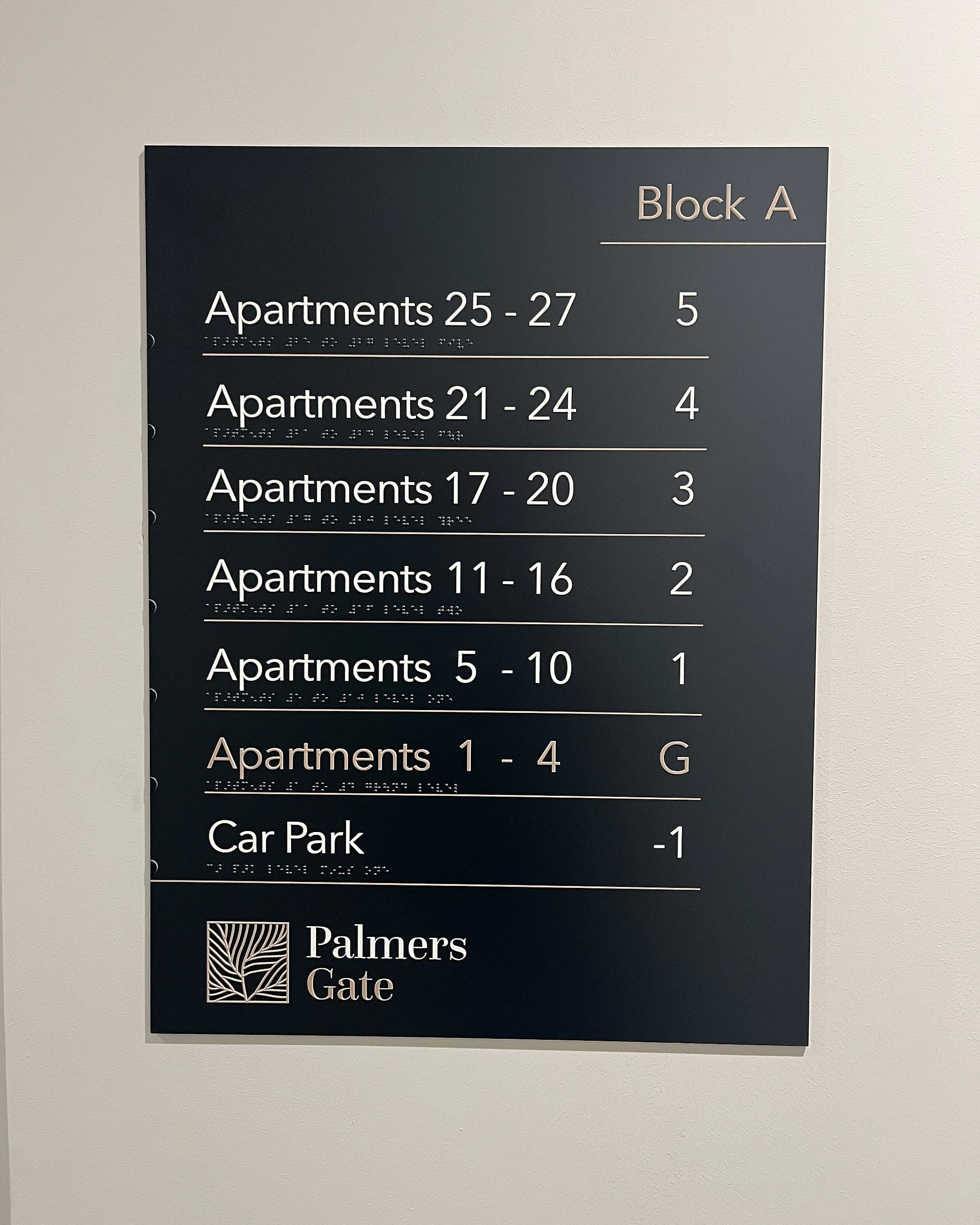 high-contrast residential floor directory at Palmers Gate: white raised lettering and braille on a near-black panel mounted on an off-white wall