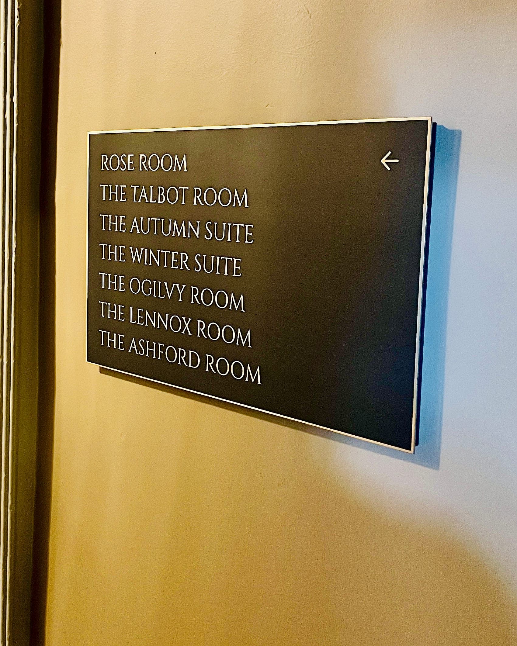 Directional wall sign at Carton House listing seven named meeting rooms and suites with a left arrow, in white lettering on a dark panel with a metal border