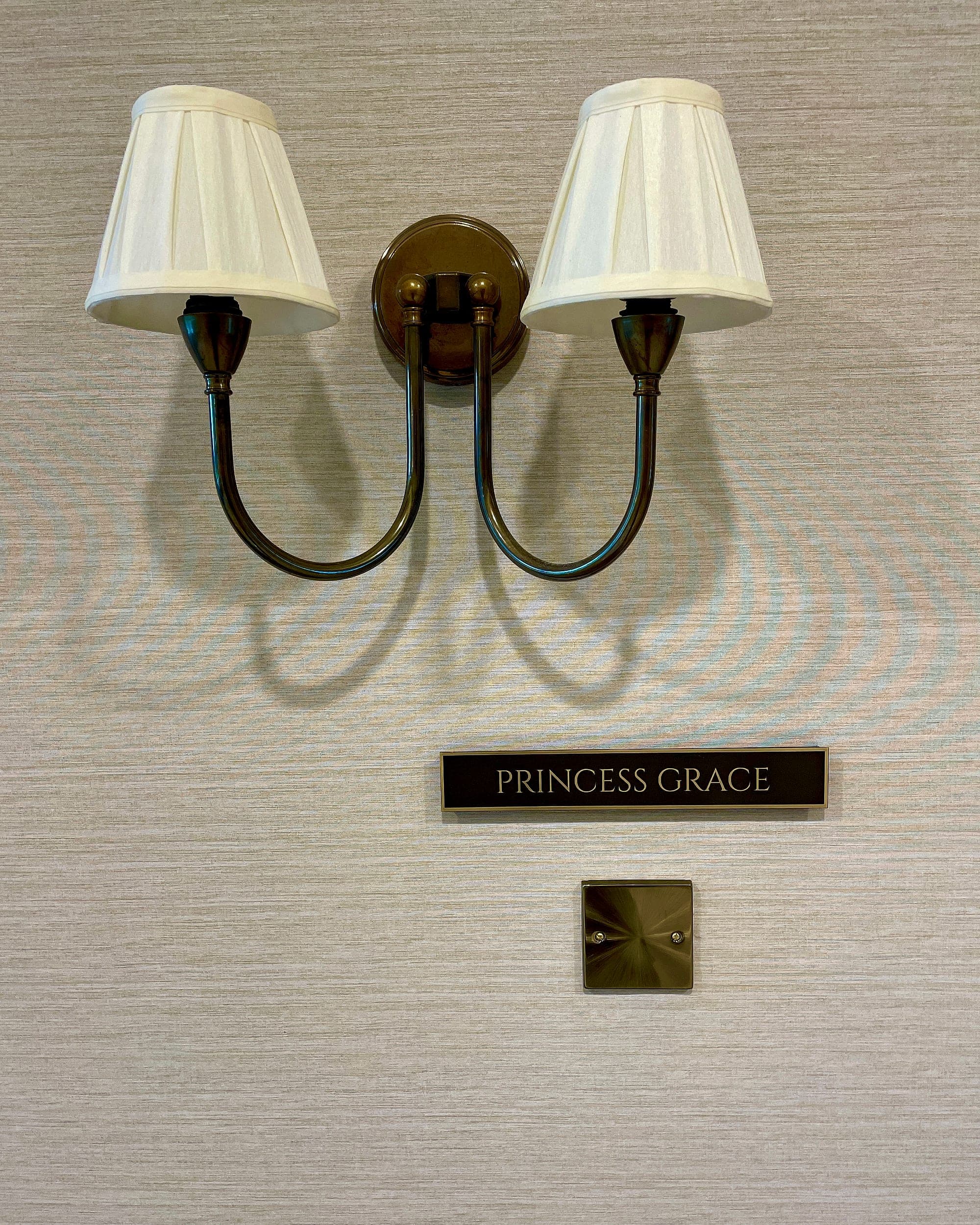 Carton House hotel signage detail: engraved brass room-name plaque reading Princess Grace, mounted beside a twin brass wall light on silk-weave wallcovering