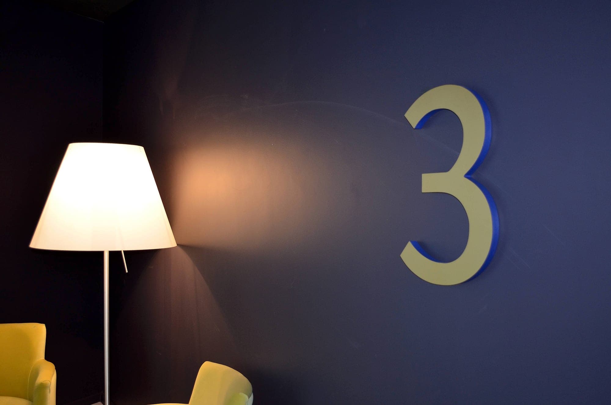 An oversized brass-finish numeral 3 mounted on a deep blue lift-lobby wall at The Marker Hotel, Dublin, beside a floor lamp and lounge chairs