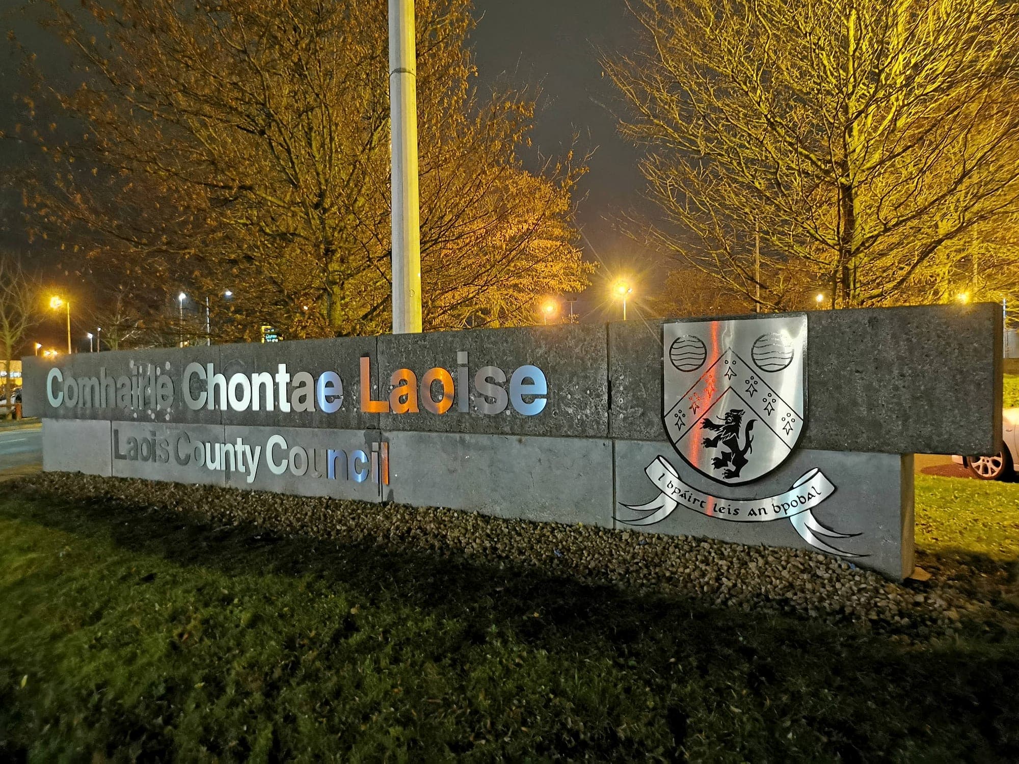 Illuminated external lettering requiring signage maintenance: bilingual Irish and English lettering with the county crest at Laois County Council's head office, on a stone-clad boundary wall, lit at night