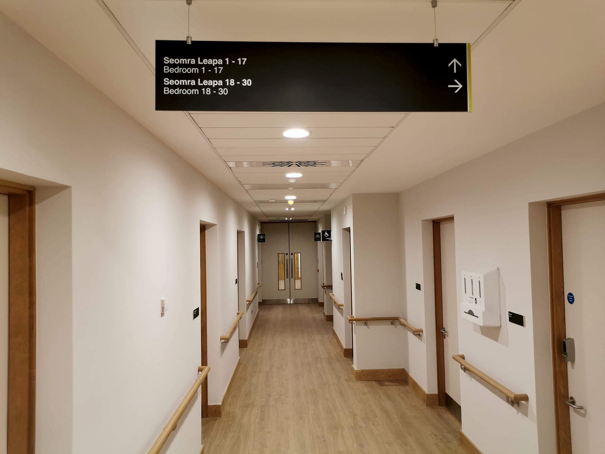 Bilingual Irish and English suspended corridor sign directing to bedroom number ranges with up and right arrows, in a healthcare corridor with handrails to both walls