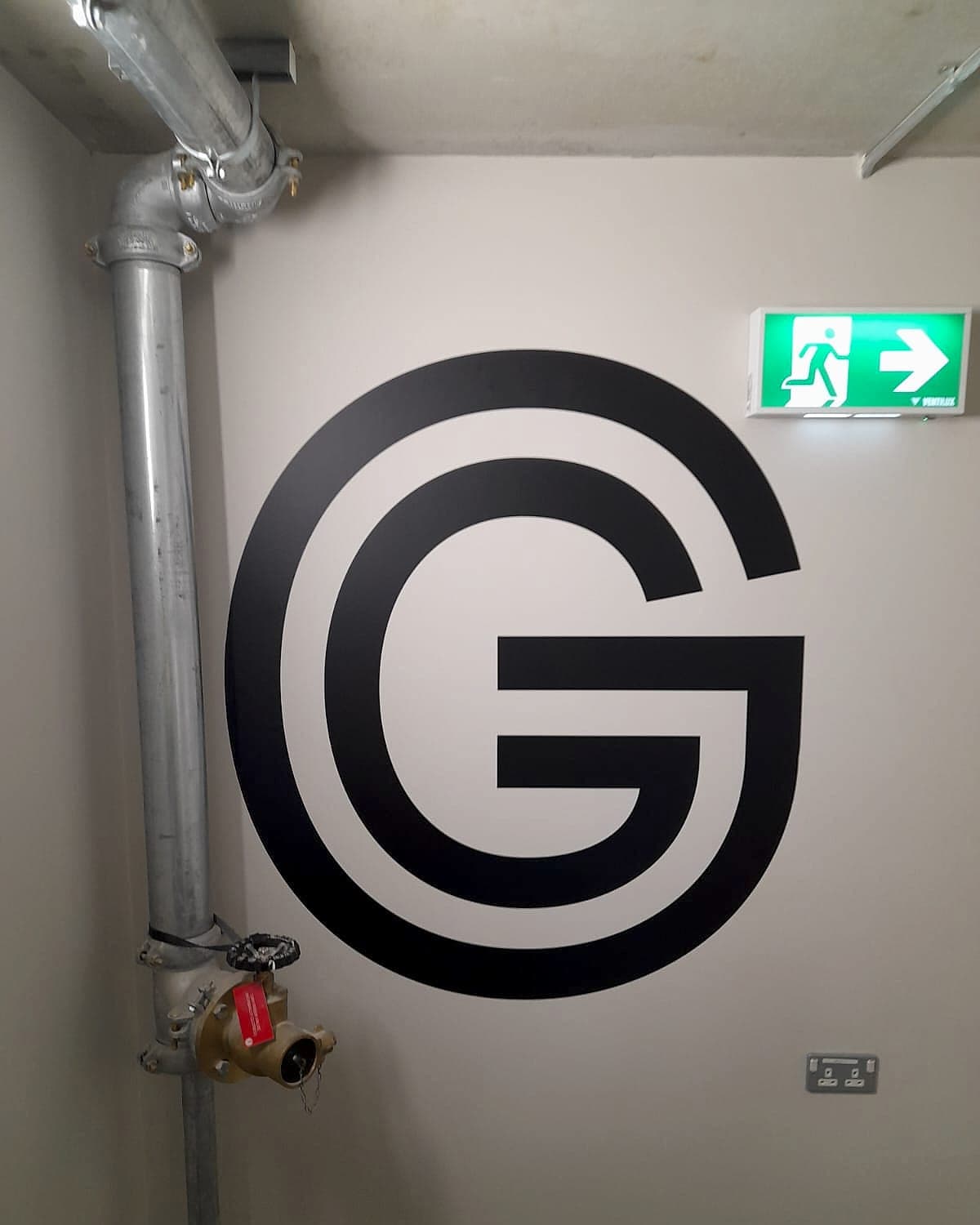 Marshal's Yard, ISO 7010 emergency exit sign: a maintained green luminaire showing the E002 running man pictogram and a directional arrow, mounted beside a large painted level letter and a dry riser inlet