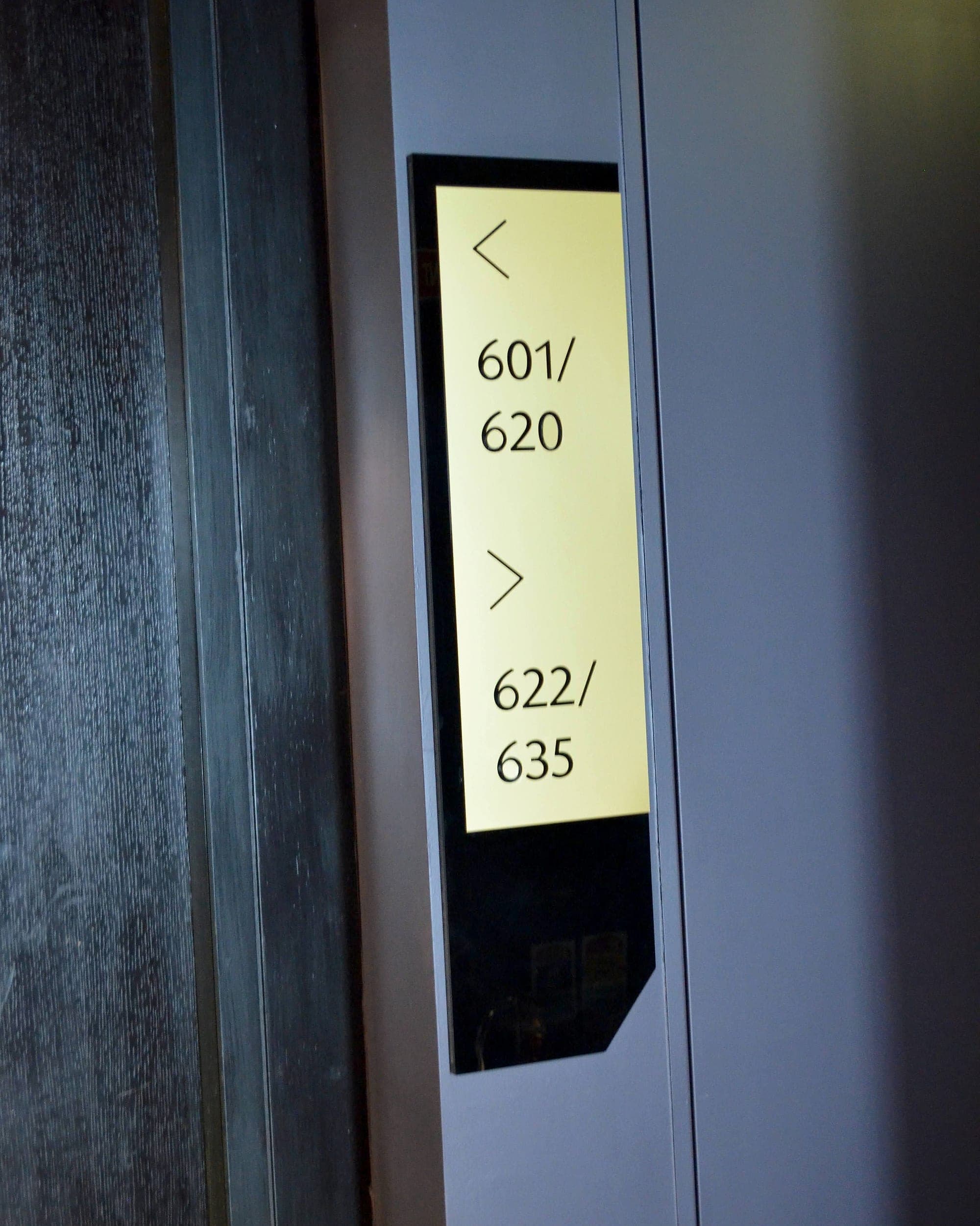 Vertical directional sign at a lift lobby in The Marker Hotel showing room number ranges 601 to 620 to the left and 622 to 635 to the right, in dark bronze with a brass-coloured face