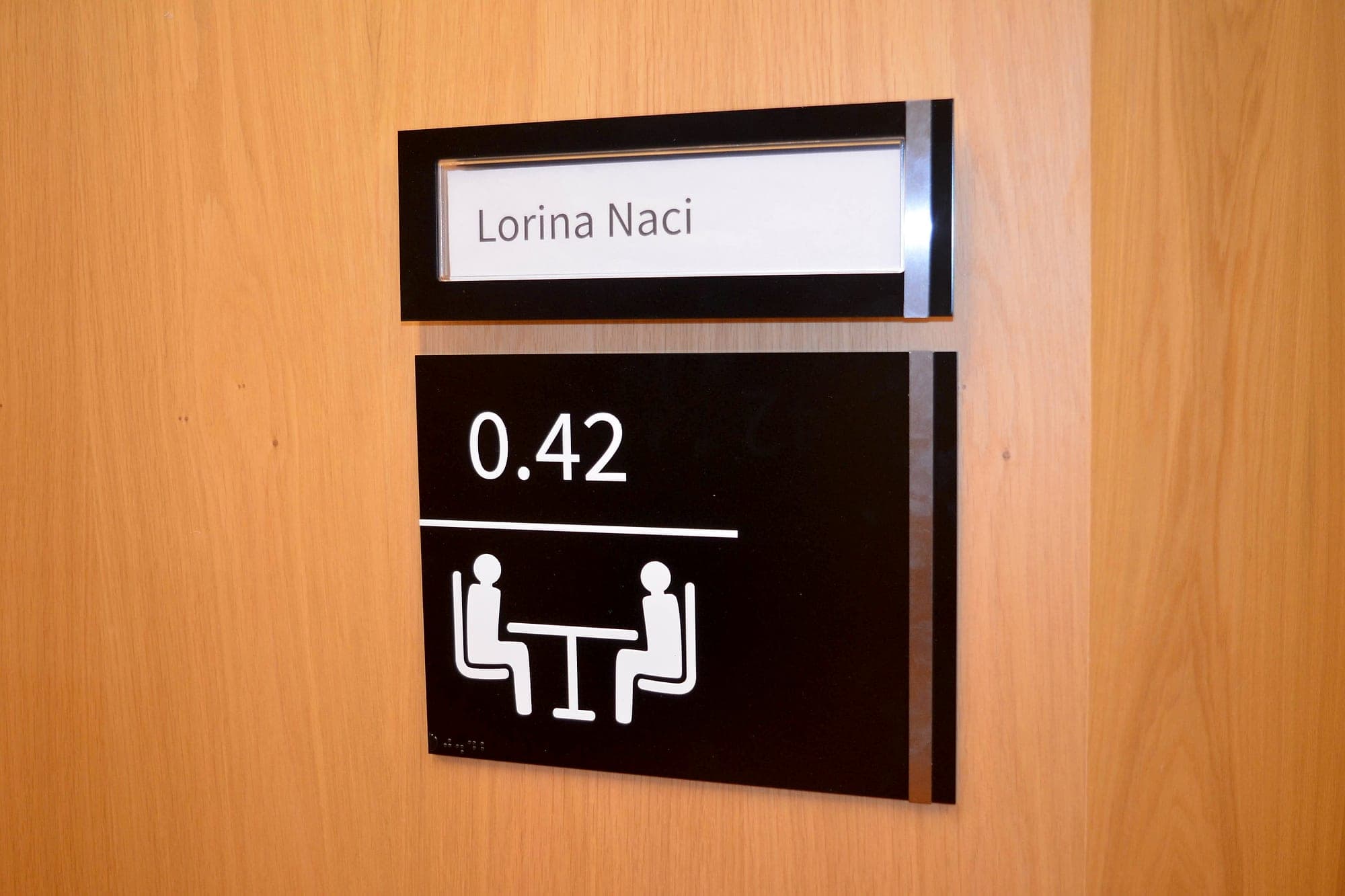The static equivalent of smart building signage at Trinity GBHI: a meeting room door sign with a fixed panel carrying room number 0.42, a two-person meeting pictogram and braille, below a slide-in insert strip holding the occupant's name