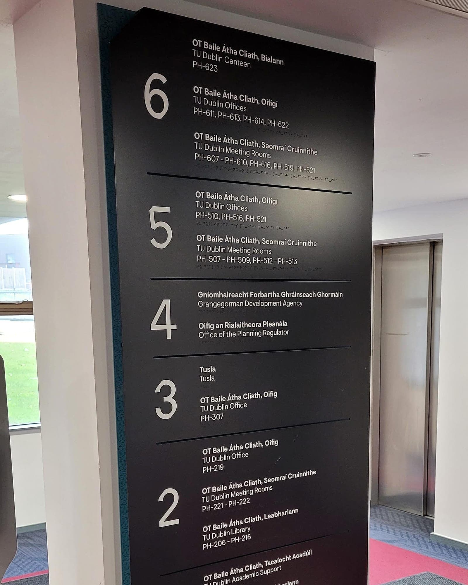 Bilingual Irish and English multi-occupier building directory at Park House, TU Dublin: a tall fabricated panel listing six levels against room-number ranges for TU Dublin, the Grangegorman Development Agency, Tusla and the Office of the Planning Regulator, with braille beneath each block