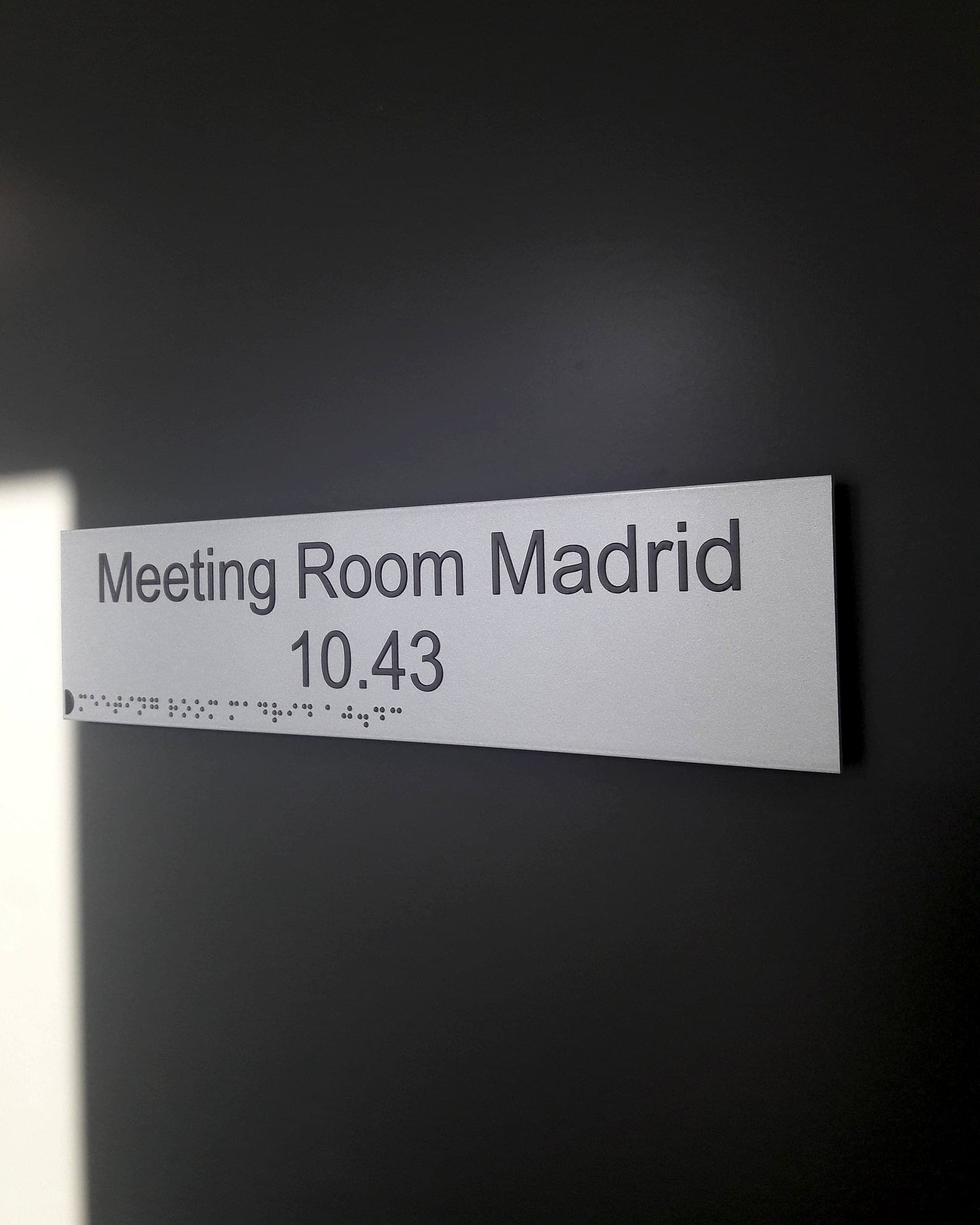 Corporate room sign reading Meeting Room Madrid 10.43 in raised tactile mixed-case lettering with braille beneath, on a brushed silver panel against a dark wall
