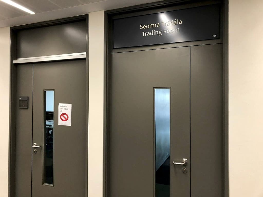 The Business School: a non-compliant printed A4 prohibition notice with an empty red circle-and-bar symbol stuck to a lab door, beside a compliant bilingual Irish and English room sign above the adjoining doors