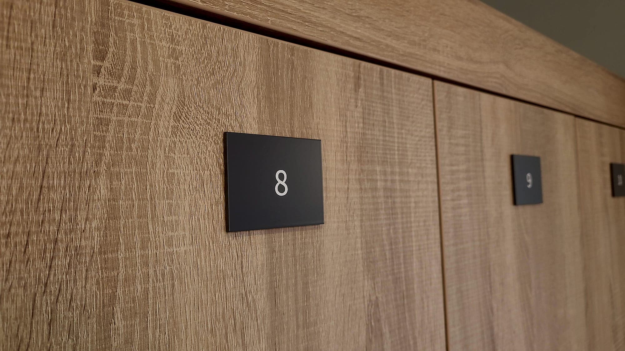 Identification stripped to a numeral: a run of oak-faced staff lockers carrying small dark plaques with white numerals only, 8 and 9, no name, function or occupant shown