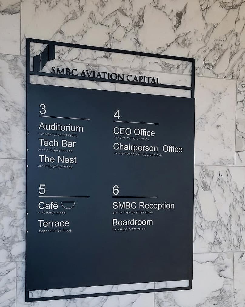 Corporate lift-lobby floor directory in office signage at SMBC: a dark tactile panel on a marble wall listing levels three to six with named spaces and braille under every line